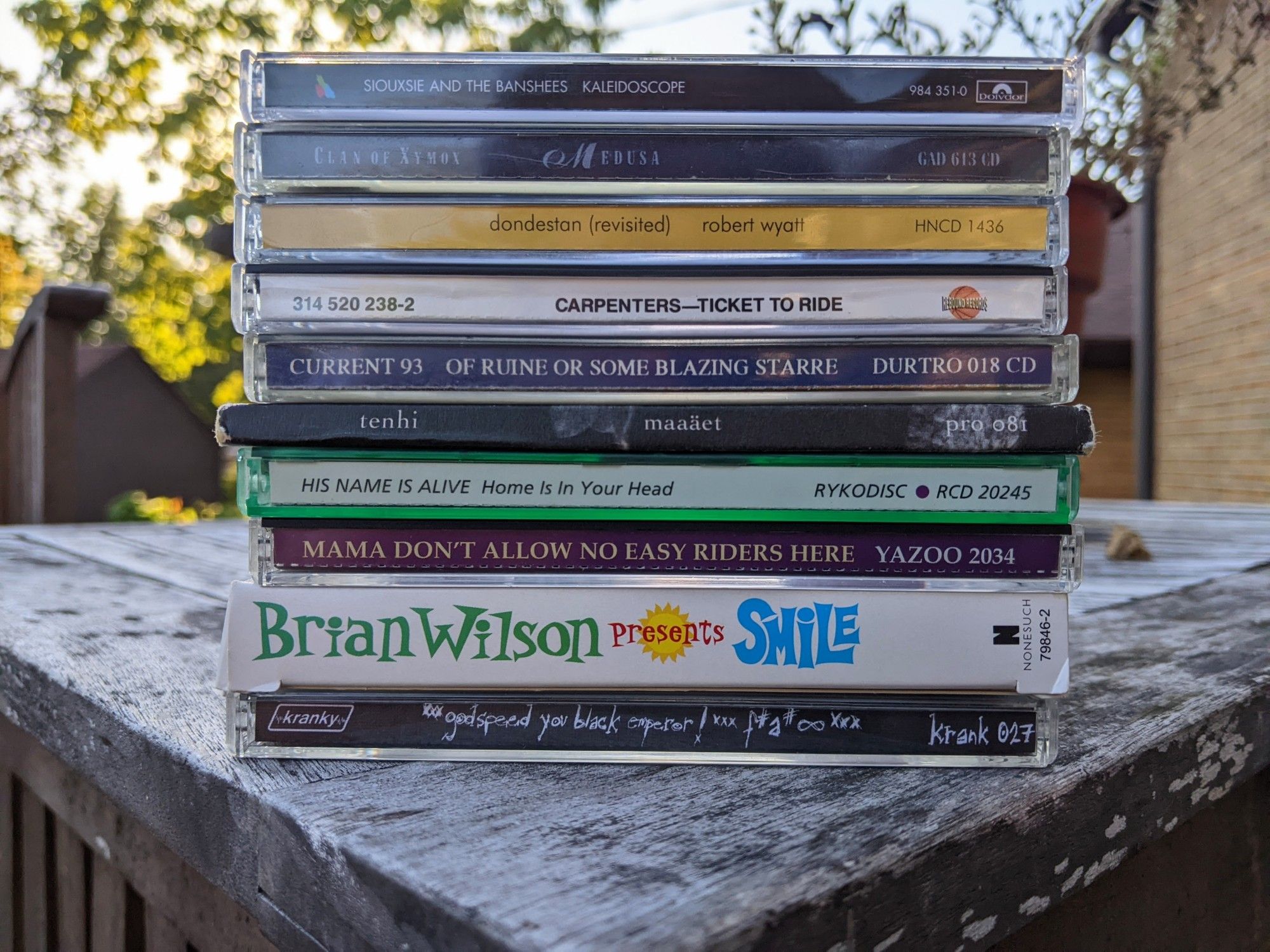 Stacked CD playlist on weathered wooden patio table, set at an approximately equilateral angle to make a triangle with the corner of the table. Autumn afternoon light.

In order played, bottom to top:

Godspeed You! Black Emperor F♯ A♯ ∞
Brian Wilson Presents SMILE
Various MAMA DON'T ALLOW NO EASY RIDERS HERE
His Name is Alive HOME IS IN YOUR HEAD
Tenhi MAAAËT
Current 93 OF RUINE OR SOME BLAZING STARRE
Carpenters TICKET TO RIDE
Robert Wyatt DONDESTAN REVISITED
Clan of Xymox MEDUSA
Siouxsie and The Banshees KALEIDOSCOPE