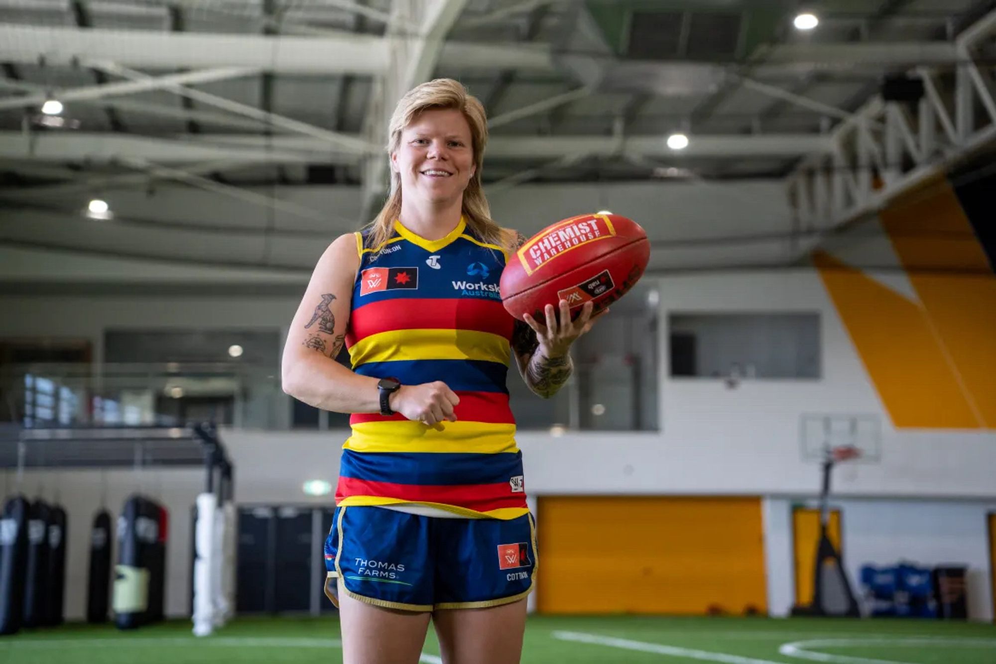 Jess Waterhouse stands holding an AFLW ball, with the other hand posed as though preparing to handball it. Her hair is shoulder length blonde hair in back and far shorter along the top and sides.