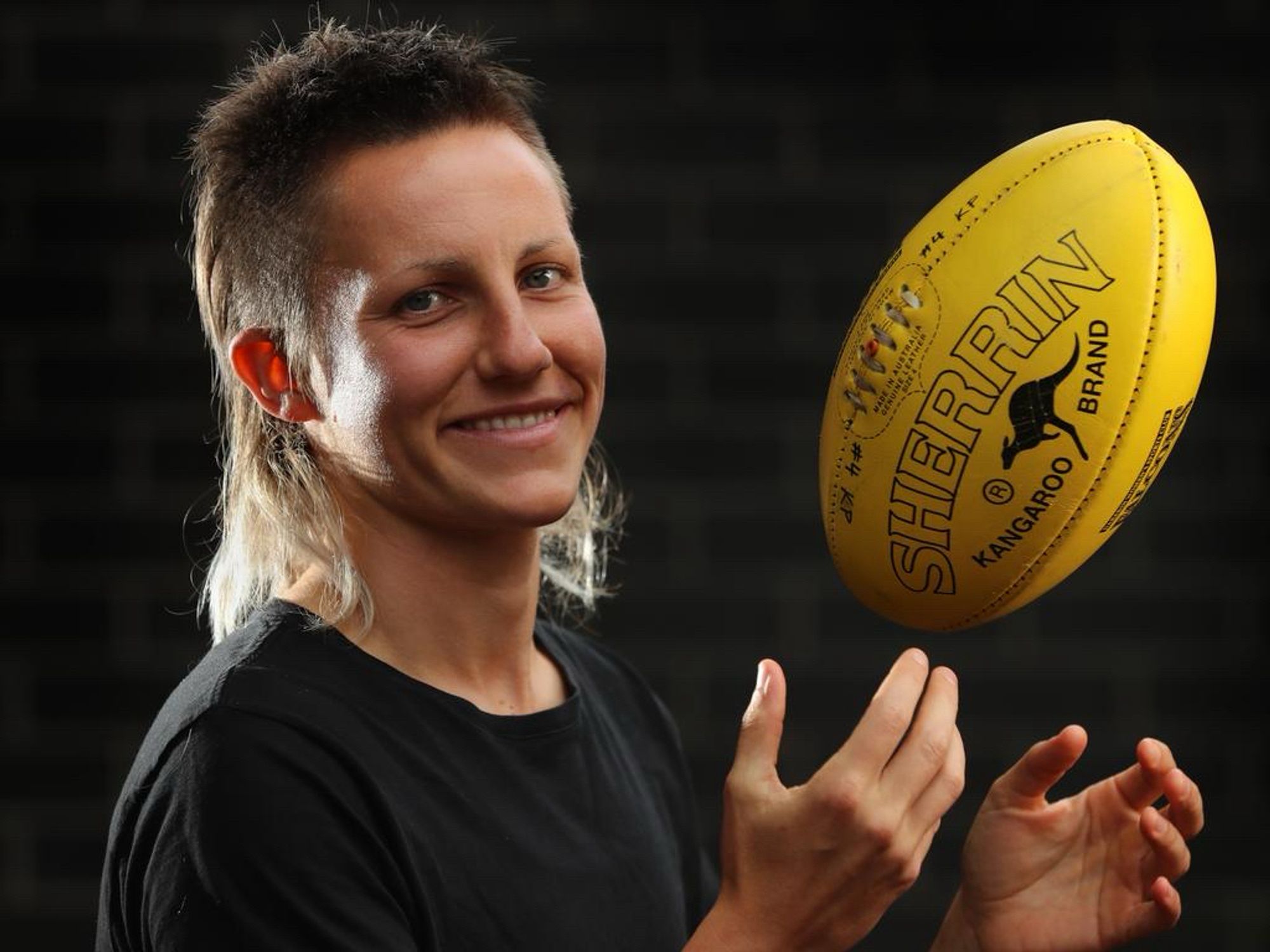 karen “paxy” paxman poses for a portrait tossing an aussie rules football. her brown hair is shorn with what looks like a #4 up top, a #2 on the side. The back of her hair, in addition to being down to her shoulder, is also dyed blonde at the tips.