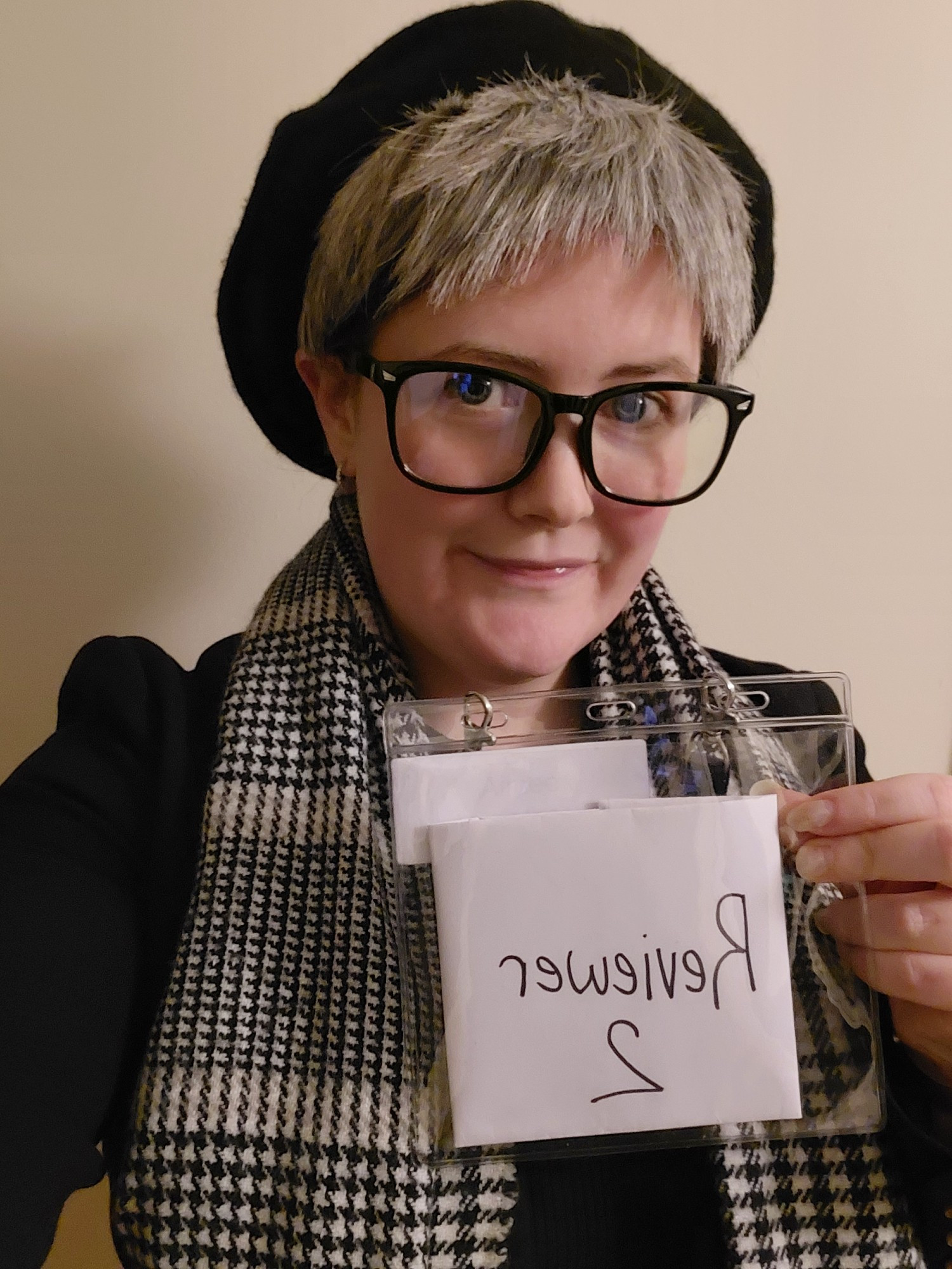 Aimee dressed up as an interpretation of Reviewer 2. She wears a lanyard that says "Reviewer 2", a grey haired wig with black beret, glases, a black blazer and black and white scarf.