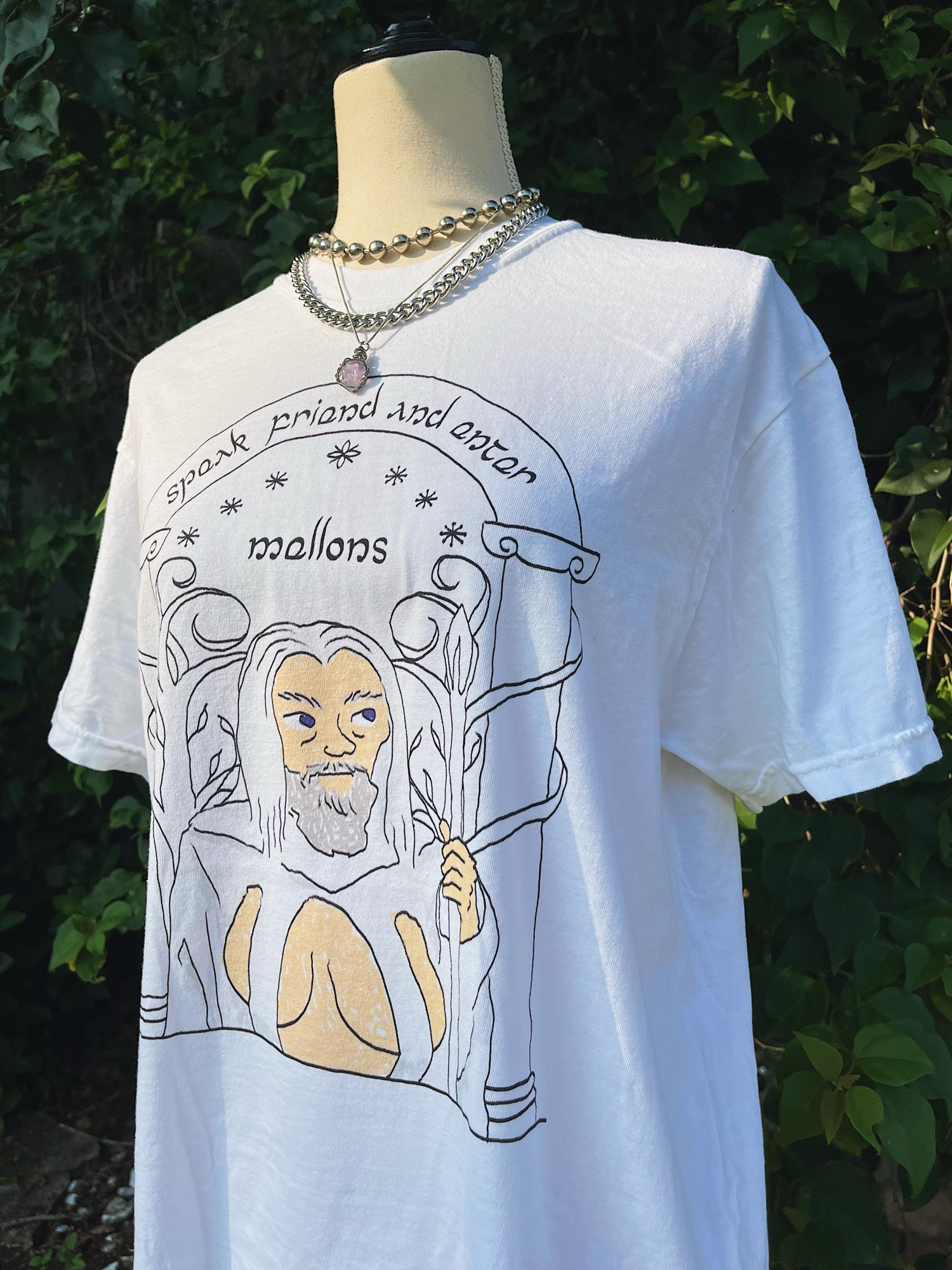 product photo of a scribble drawing of Gandalf the White, the wizard from Lord of the Rings. He’s looking off to the right, holding a staff, and is wearing a sexy strappy shirt that shows off boobs. He’s standing in front of the Doors of Durin. A door with curly tree branch adornments and text that says, “speak friend and enter: mellons” in half elvish font.