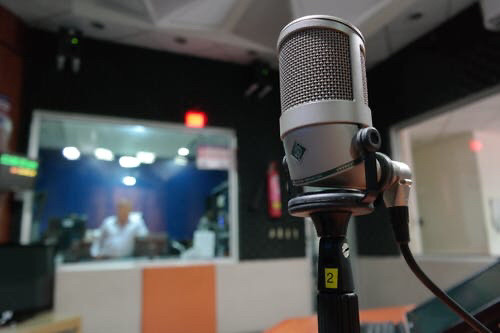 A Neumann microphone positioned in a recording studio. A window into the engineer’s booth is visible with a director watching.