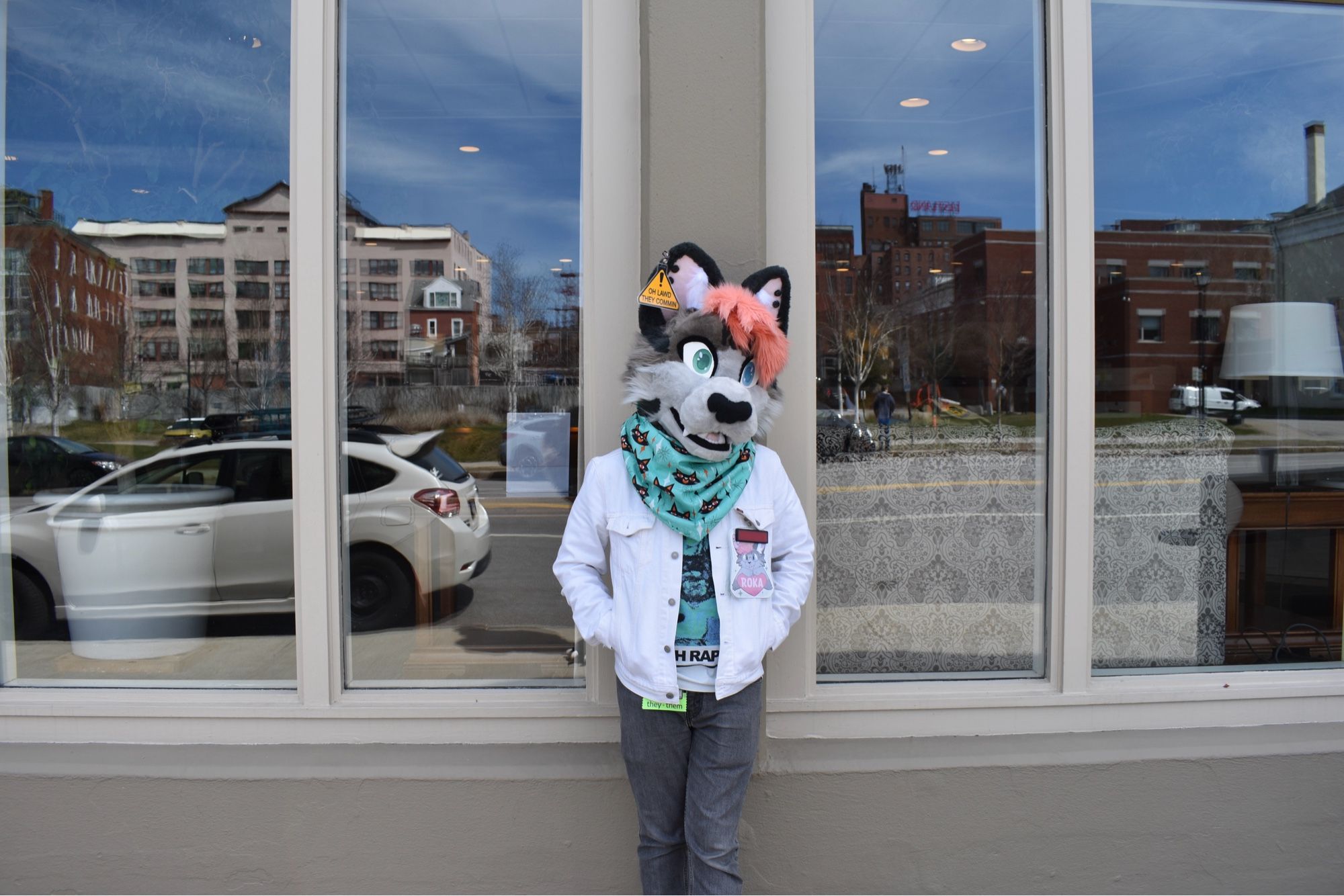 A photo taken outside. I'm leaning against a beige building with windows at either side, looking at the camera with my head tilted a bit. 
I'm wearing my fursuit head of my sona Roka, a gray cat with a light gray face, two-tone pink hair, and a heart-shaped nose. I've got a teal bandana with little cats on it around my neck. I'm wearing a white jacket, a george clanton t-shirt, and gray jeans.