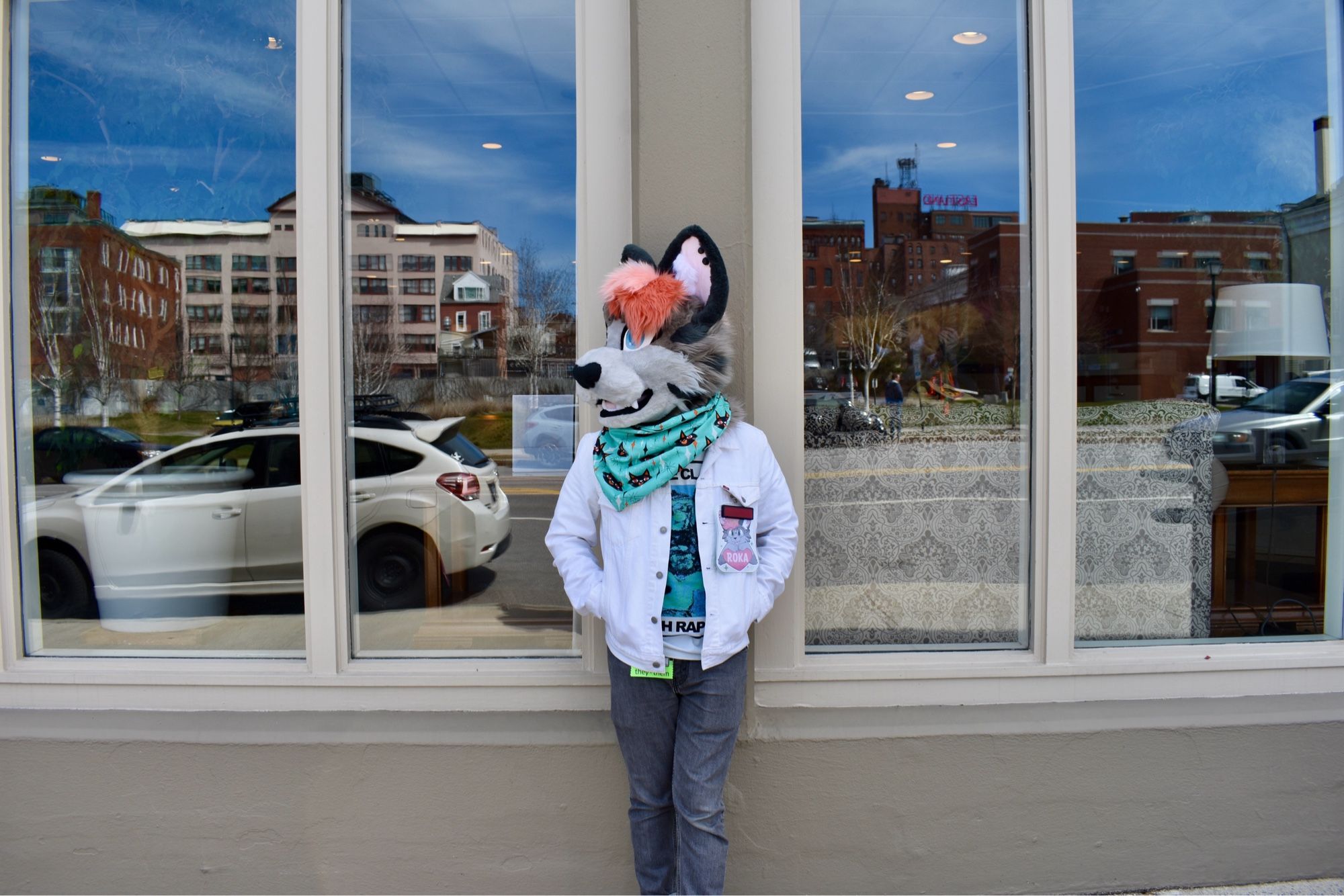 A photo taken outside. I'm leaning against a beige building and looking to the left off-camera, with windows at either side. 
I'm wearing my fursuit head of my sona Roka, a gray cat with a light gray face, two-tone pink hair, and a heart-shaped nose. I've got a teal bandana with little cats on it around my neck. I'm wearing a white jacket, a george clanton t-shirt, and gray jeans.