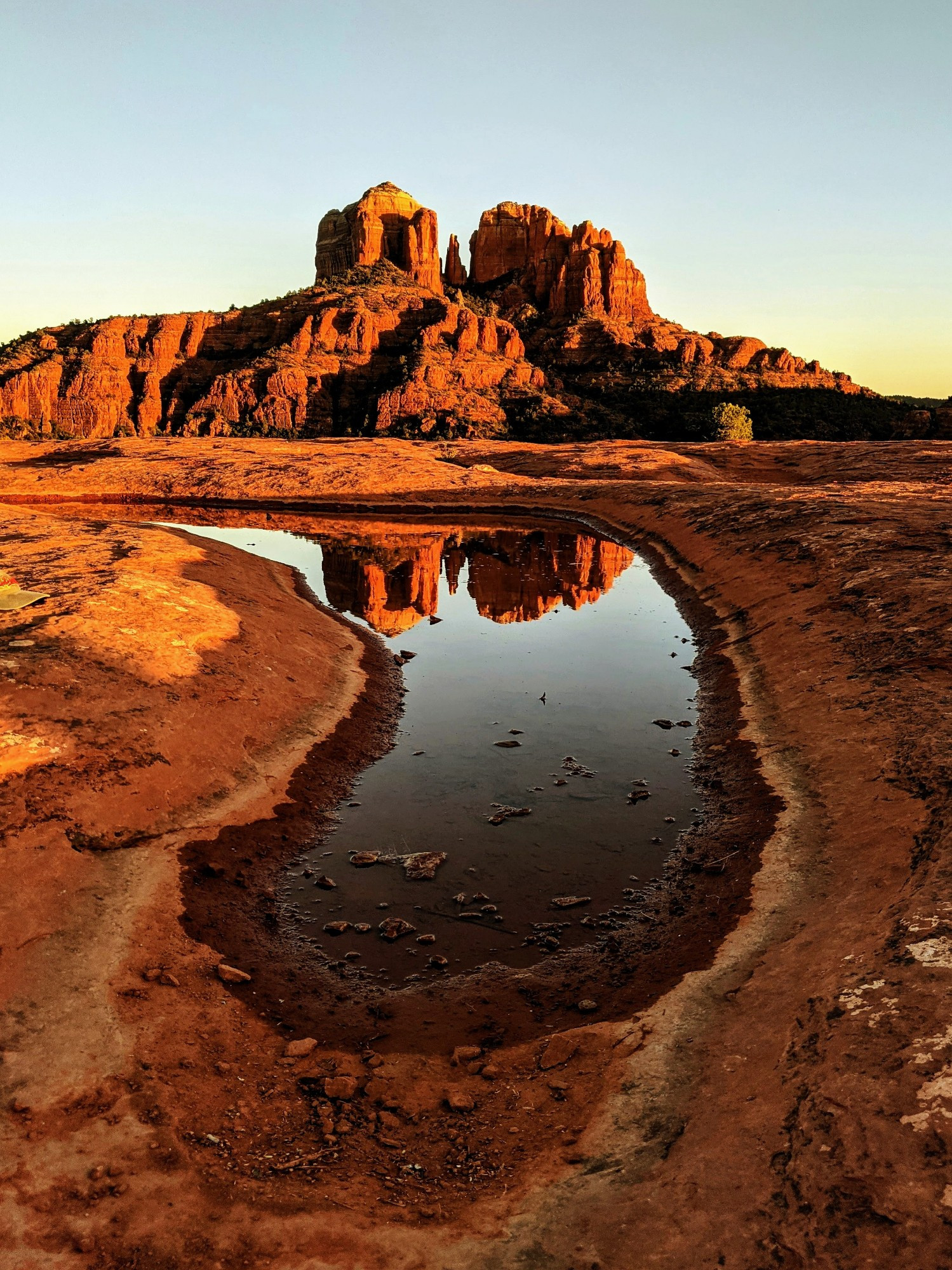 Cathedral Rock in Sedona reflected in a large curved puddle as the sun sets. The rocks are glowing orange and the puddle looks like mercury. The beautiful buttes are set against a pale, blue sky with a bit of bright yellow on the right side. 