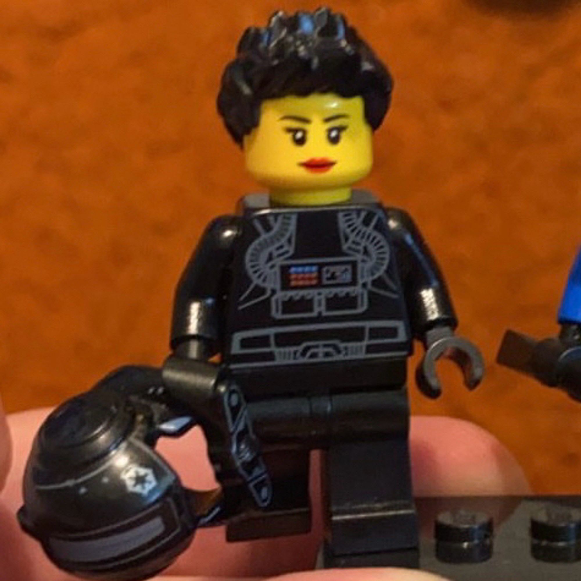 A Lego minifigure with plain black legs and a black torso with flight suit details, holding a black pilot helmet. She has a yellow skin tone with red lipstick and black hair in a spiked style.