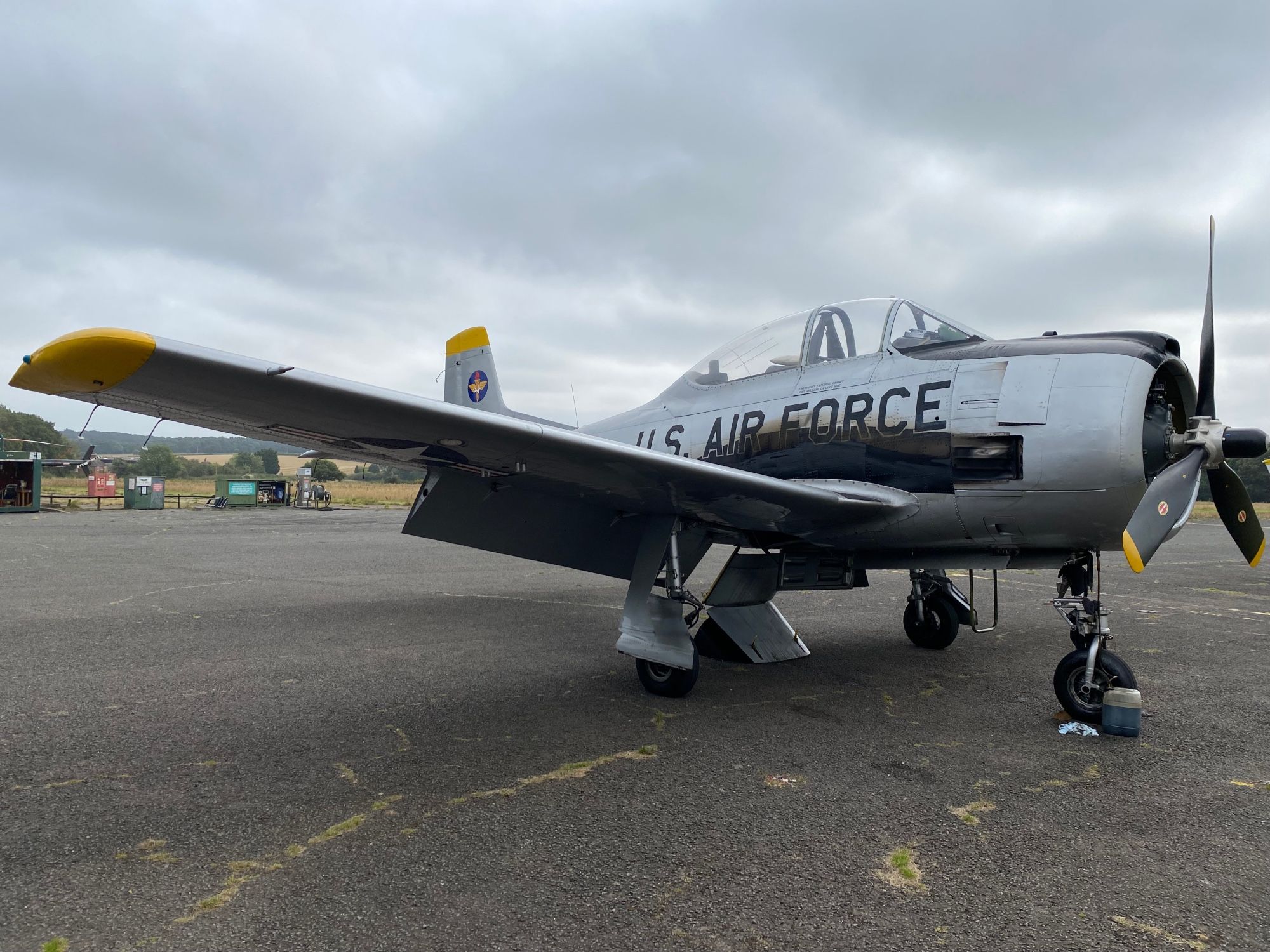 T-28 Trojan aeroplane in US air force markings sitting on the apron of an airfield. The silver aeroplane chimes with the thundery grey skies and deserted setting. There’s a feeling on uneasiness in the air.