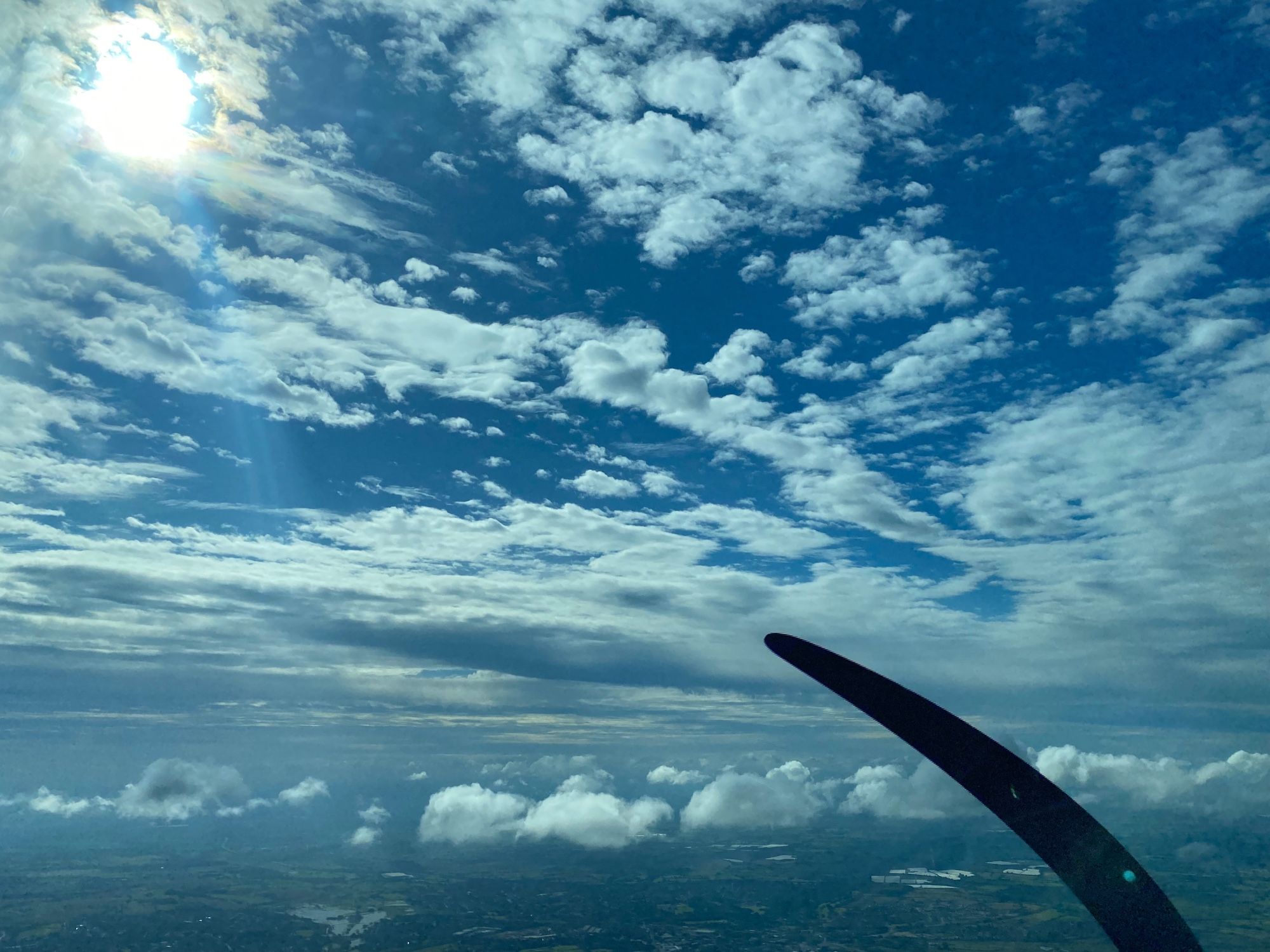 High level cloud stipples a deep blue sky, partially obscuring the sun. A lower level line of small puffs of cumulus clouds bobs above the ground. A curved black propeller blade cuts across the bottom right quadrant of the picture. It’s a picture of joy in the air.