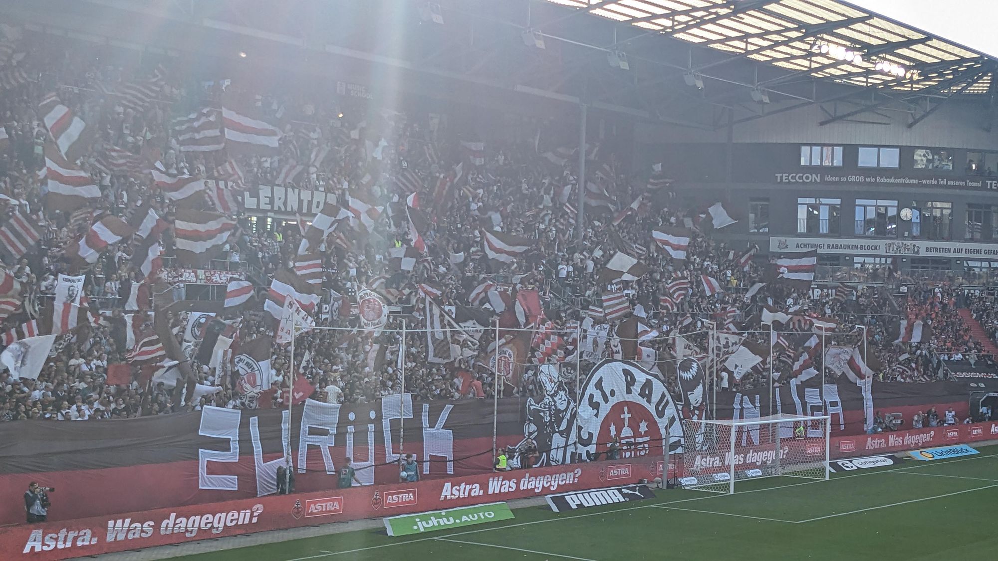 Bildbeschreibung von ChatGPT:
Das Foto zeigt eine lebhafte Szene aus einem Fußballstadion während eines Spiels. Im Vordergrund ist das Spielfeld mit einem Tor zu sehen, während der Fokus auf den voll besetzten Fanblock der Zuschauer liegt. 

Die Fans im Stadion, wahrscheinlich Anhänger des FC St. Pauli, schwenken zahlreiche Fahnen in den Vereinsfarben Braun, Weiß und Rot. Einige Fans halten große Banner hoch, die zusammengenommen das Wort "ZURÜCK" bilden. Der Fanblock ist sehr dicht gefüllt, und die Stimmung scheint ausgelassen und energiegeladen zu sein.

Im Hintergrund sieht man die Tribüne mit mehreren Werbebannern, auf denen unter anderem „Astra. Was dagegen?“ zu lesen ist, sowie eine Anzeigetafel und einige Gebäudestrukturen des Stadions. Der Fanblock ist mit einem großen Transparent geschmückt, auf dem das Logo des FC St. Pauli mit dem markanten Totenkopf zu sehen ist. 

Die Atmosphäre im Stadion wirkt sehr mitreißend und zeigt die Leidenschaft der Fans während des Spiels.