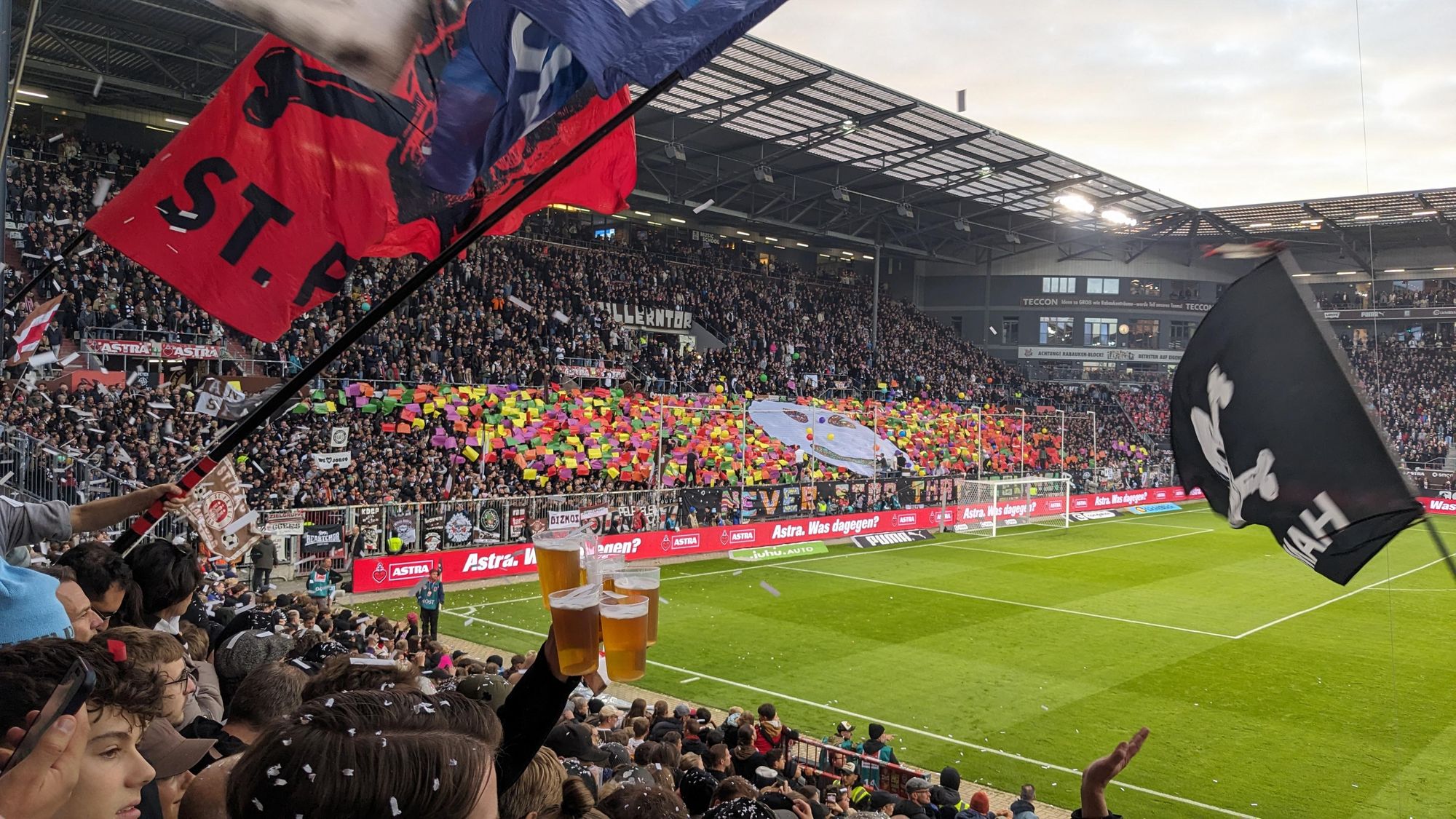 Bildbeschreibung von ChatGPT:
Dieses Foto zeigt eine lebhafte Szene aus einem Fußballstadion, höchstwahrscheinlich im Millerntor-Stadion des FC St. Pauli. Im Vordergrund schwenken Fans große Fahnen, darunter eine rote Fahne mit der Aufschrift "St. Pauli" und eine schwarze Fahne mit einem Totenkopf, dem bekannten Symbol des Vereins. Die Tribünen sind voll besetzt, und auf der gegenüberliegenden Seite der Fans gibt es eine bunte Choreografie mit farbigen Karten, die von den Fans hochgehalten werden. Im Stadion fliegt Konfetti, und es herrscht eine ausgelassene, feierliche Atmosphäre. Die Zuschauer wirken begeistert, und ein Fan hält drei Becher Bier in die Luft. Die Werbung für "Astra" am Spielfeldrand deutet auf die lokale Biermarke hin, die eng mit dem Verein verbunden ist.