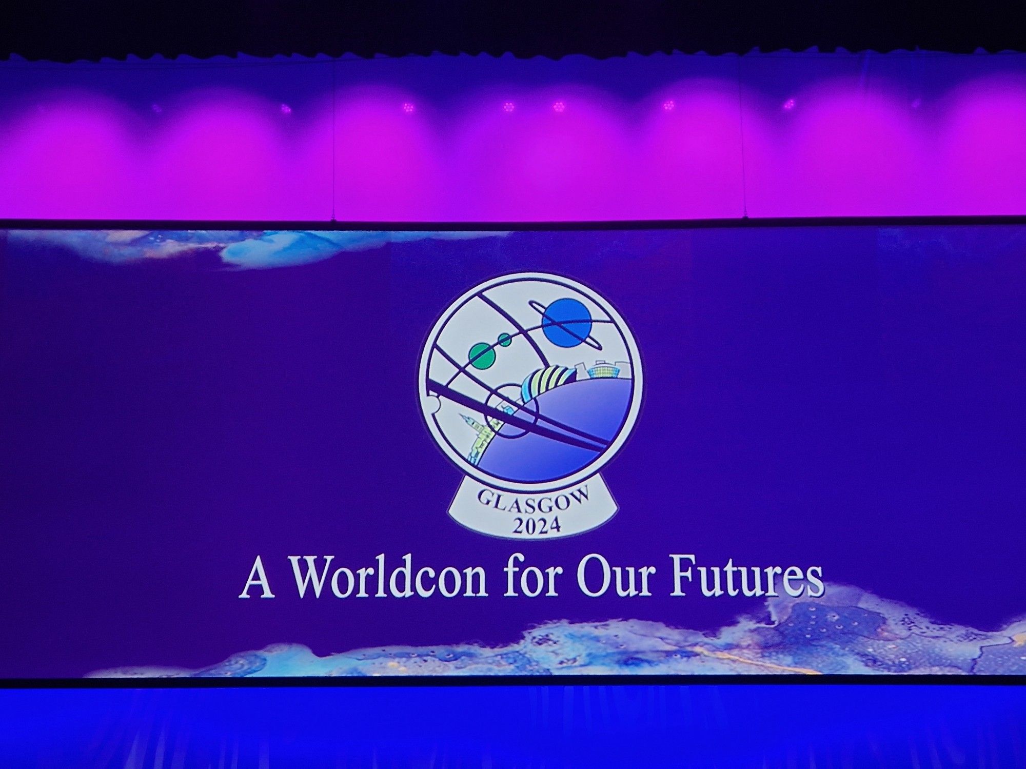 ID: Shown is a photograph of a large screen with the Glasgow 2024 logo on it and the words "A Worldcon for Our Futures". With purple and blue lights. END ID