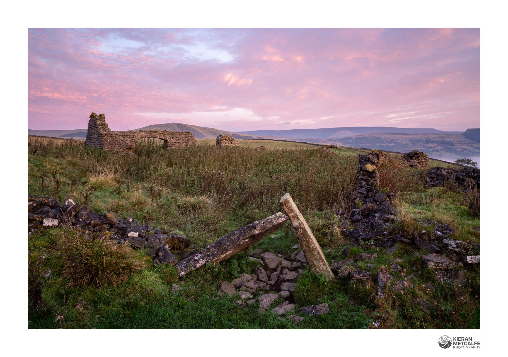 Sunrise at the remains of an old barn in the peak district. High clouds are catching a soft pink glow. A pair of old gate posts have collapsed to lean against each other in the foreground.