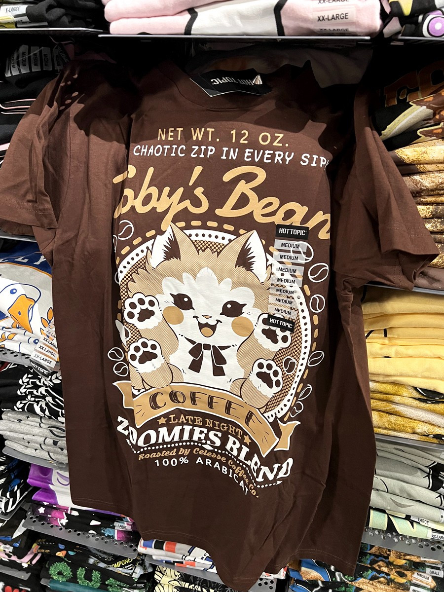 A photo of a brown t-shirt hung on a wall of shelves of stacked shirts, the print featuring a faux logo for Toby's Beans Coffee with a kitten showing off his toe beans