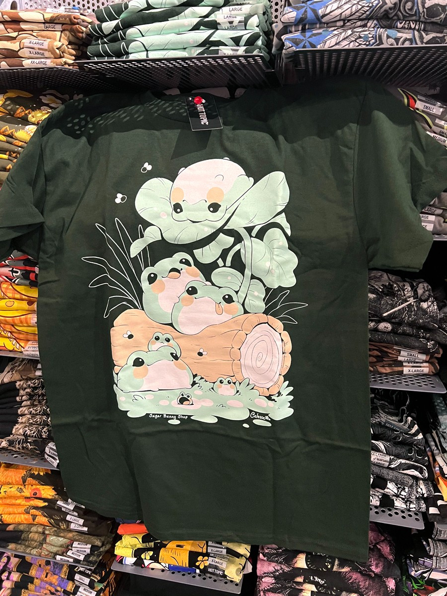 A photo of a forest green t-shirt hung on a wall of shelves of stacked shirts, the print featuring a scene of fat blob frogs piled on a log together