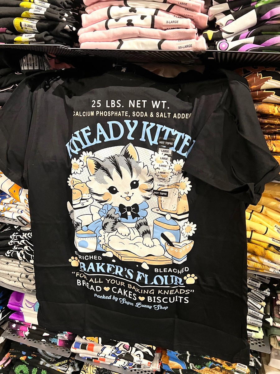 A photo of a black t-shirt hung on a wall of shelves of stacked shirts, the print featuring a faux logo for Kneady Kitten Baker's Flour with a kitten kneading dough