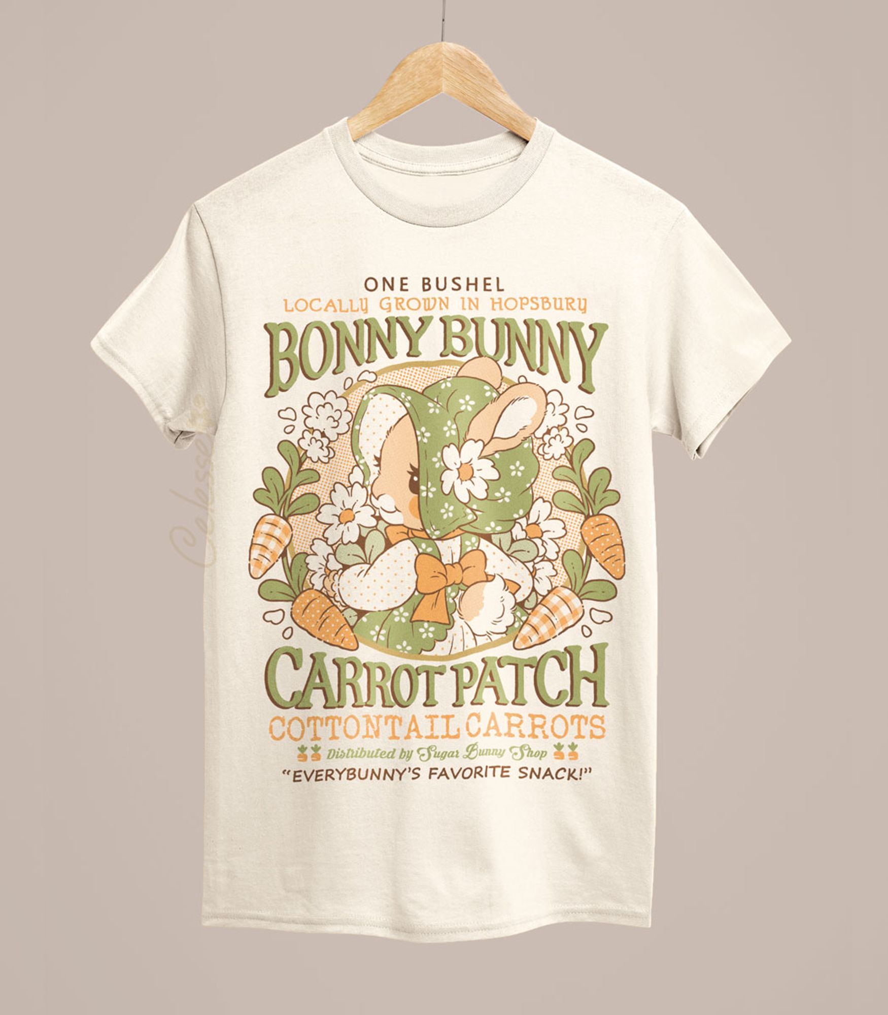 A photo of a natural color shirt on a wood hanger, the printed design features a vintage-style logo for Bonny Bunny Carrot Patch with a bunny in a floral bonnet and dress, holding a bundle of flowers and wreathed by carrots