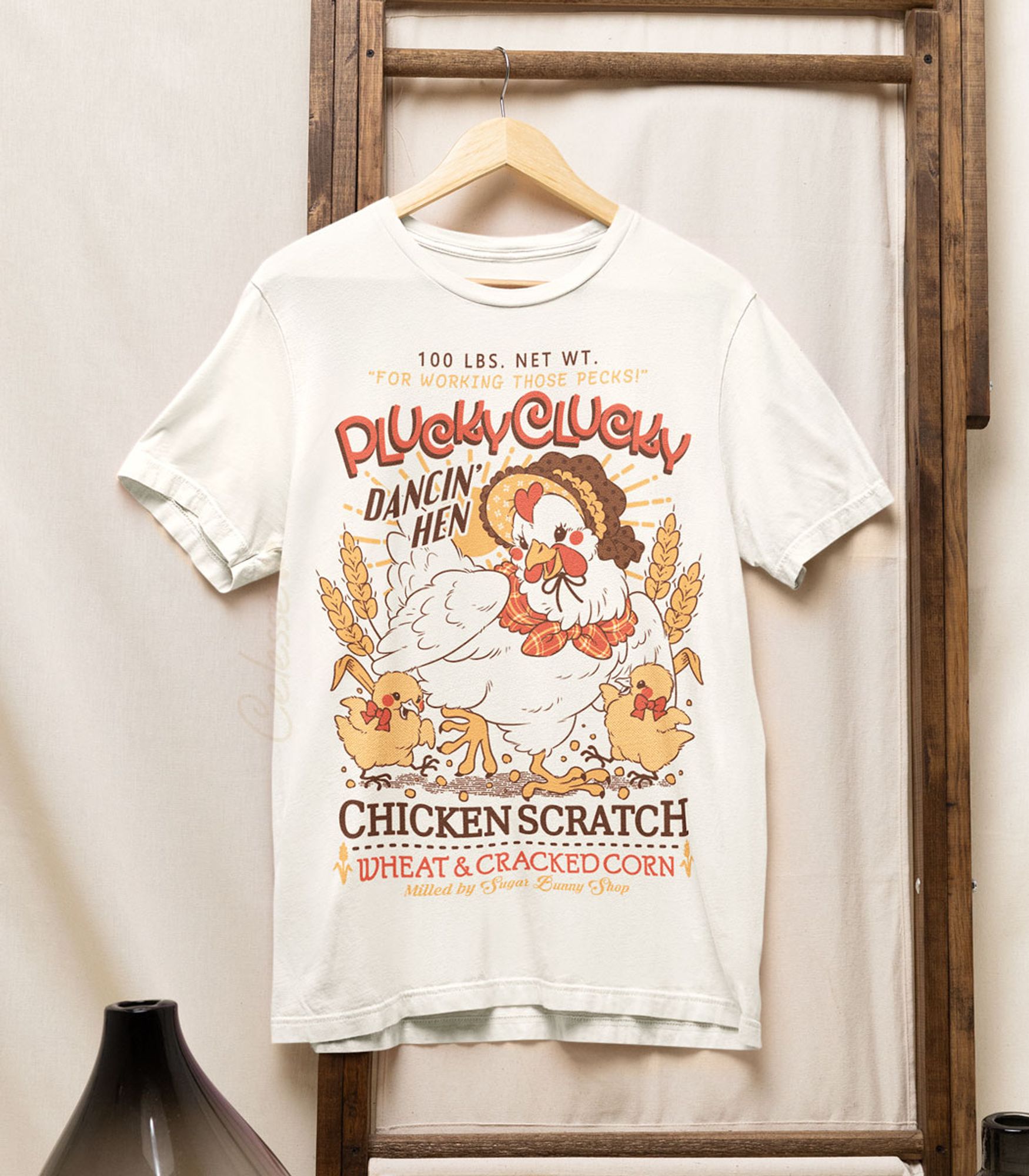 A photo of an off-white shirt on a wood hanger, hung from a wooden frame, the printed design features a vintage-style logo for Plucky Clucky Dancin' Hen Chicken Scratch with a mother hen in a bonnet dancing with two yellow chicks on either side of her