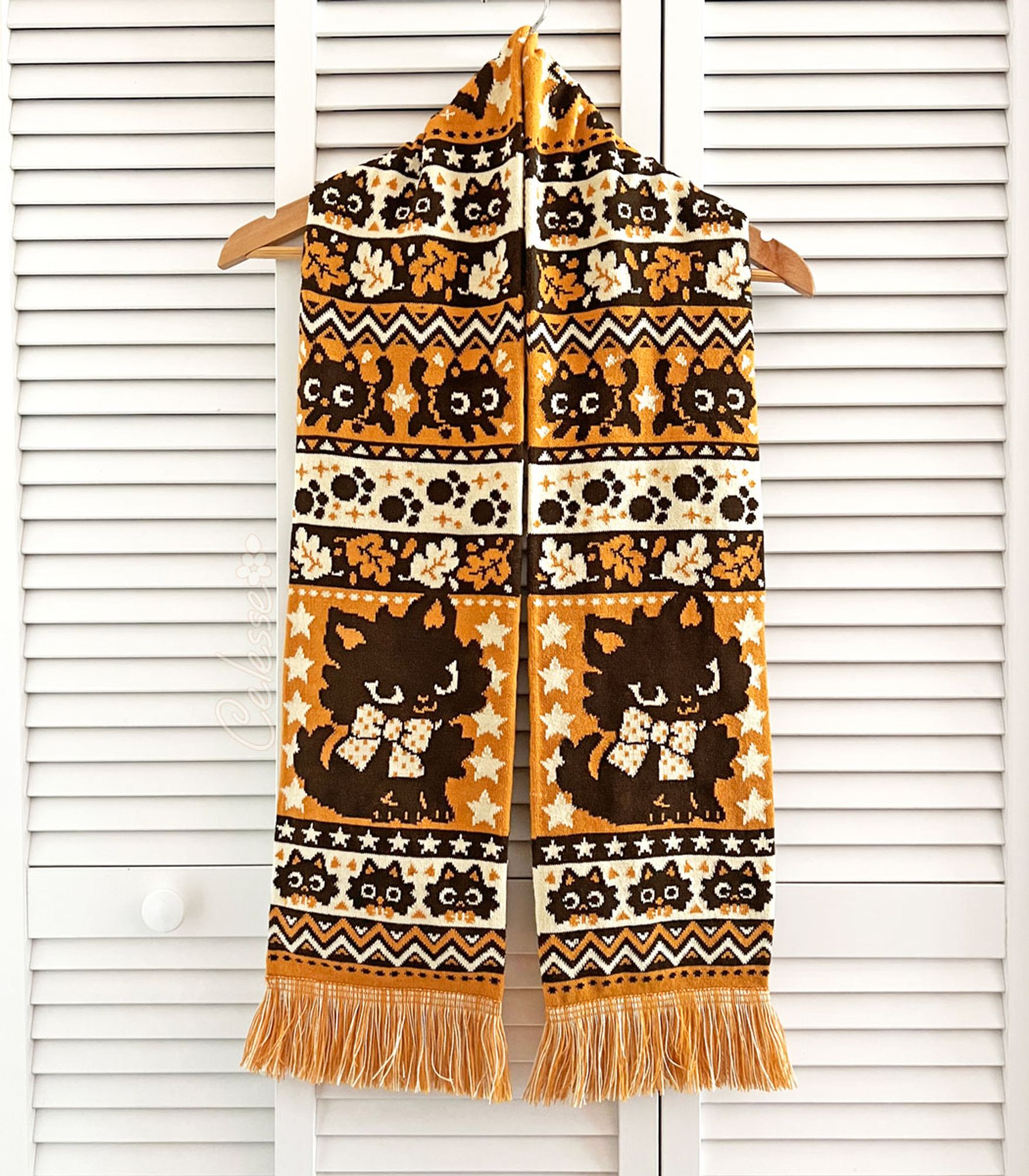 A photo of a Halloween-themed knit scarf on a wood hanger, hung on a set of white slat-walled doors. The scarf is orange, light yellow and dark brown and features rows of black cats, leaves, paw prints and other spooky motifs