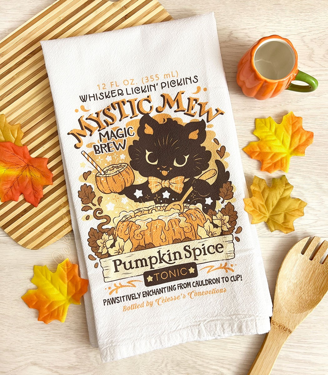 A photo of white tea towel with a print featuring a logo for Mystic Mew Pumpkin Spice Tonic, a black kitten stirring a pumpkin cauldron full of magic brew popping with stars and sparkles and holding up a cup-sized pumpkin with a straw in it. The towel lays on a table with a striped cutting board, autumn leaves, a slotted spoon and a small pumpkin-shaped mug.