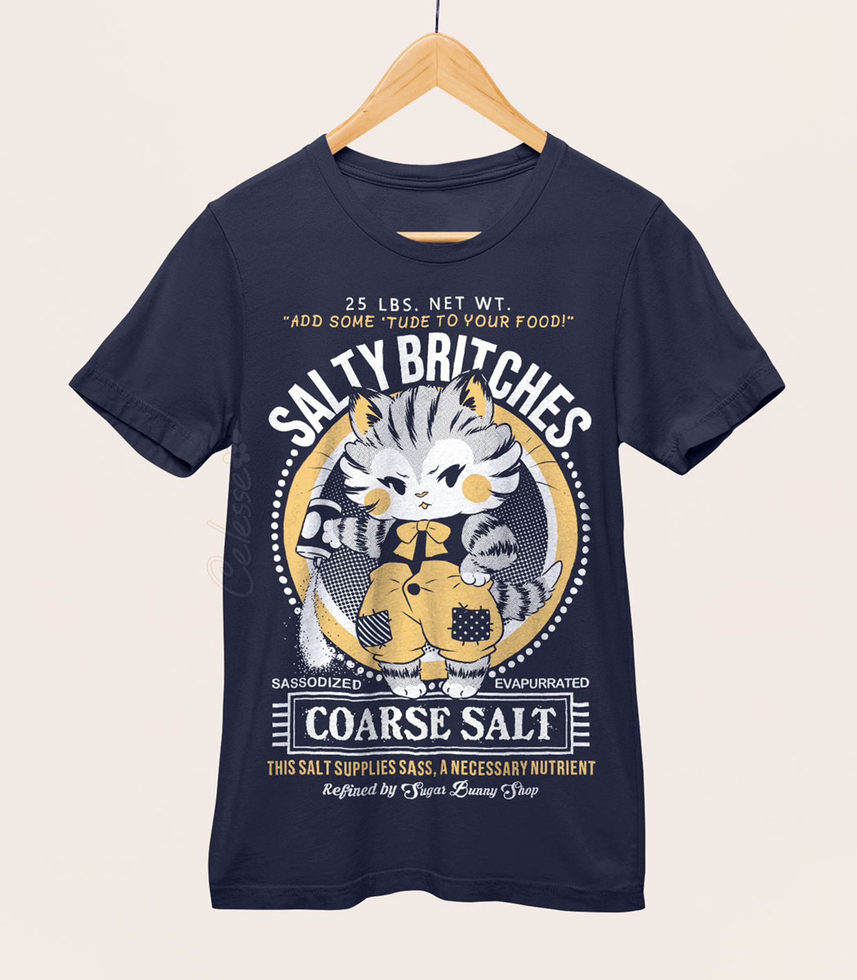 A photo of a navy blue shirt on a wood hanger, the printed design features a vintage-style logo for Salty Britches Coarse Salt with a kitten wearing patchwork britches and petulantly pouring out a container of salt