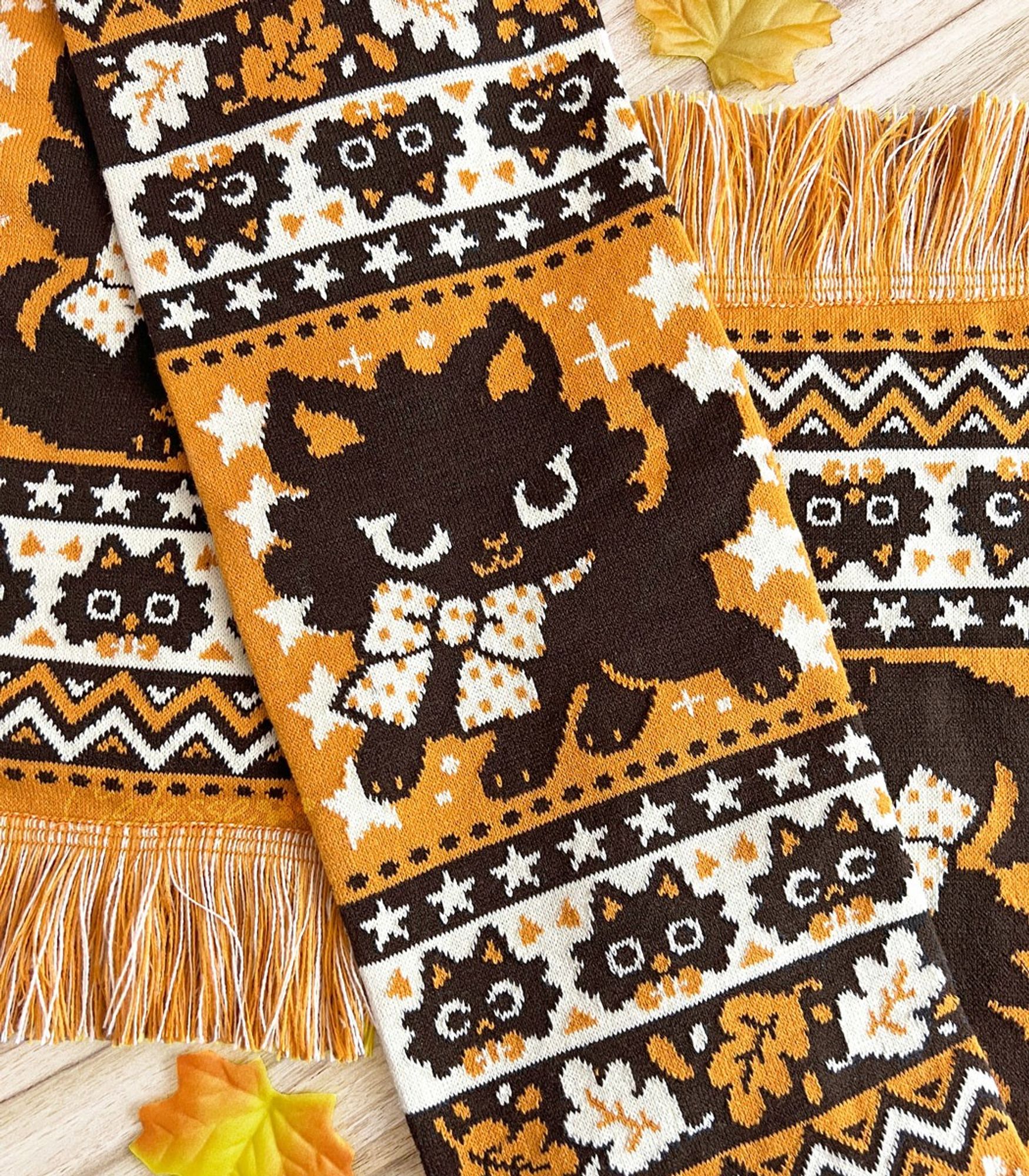 A close-up photo of a Halloween-themed knit scarf made of orange, dark brown and pale yellow yarn. A black kitten with a yellow polka dot bow poses amidst rows of stars, dots, leaves and other spooky motifs knit into the design.