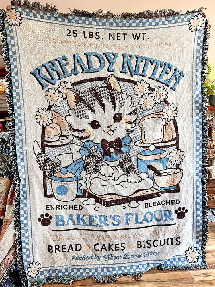 A photo of a woven throw blanket being held up. The blanket features a vintage logo design for Kneady Kitten Baker's Flour with a kitten in an apron kneading dough with bread and baking supplies around her