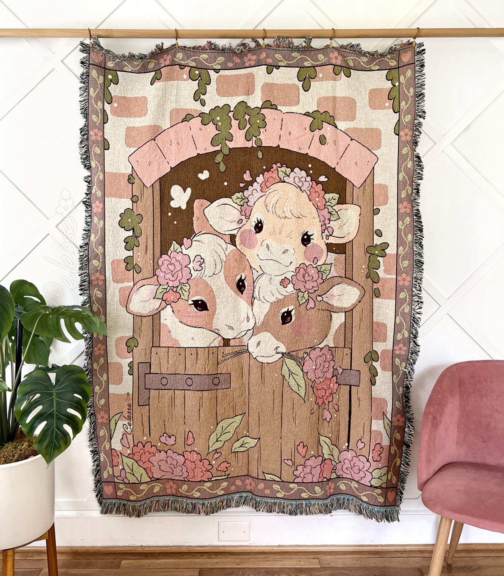 A photo of a woven tapestry blanket hung from a wooden pole against a white wall, a house plant and chair on either side of it. The blanket features 3 country cows looking through a barn door set in a brick wall decorated with flowers and foliage.