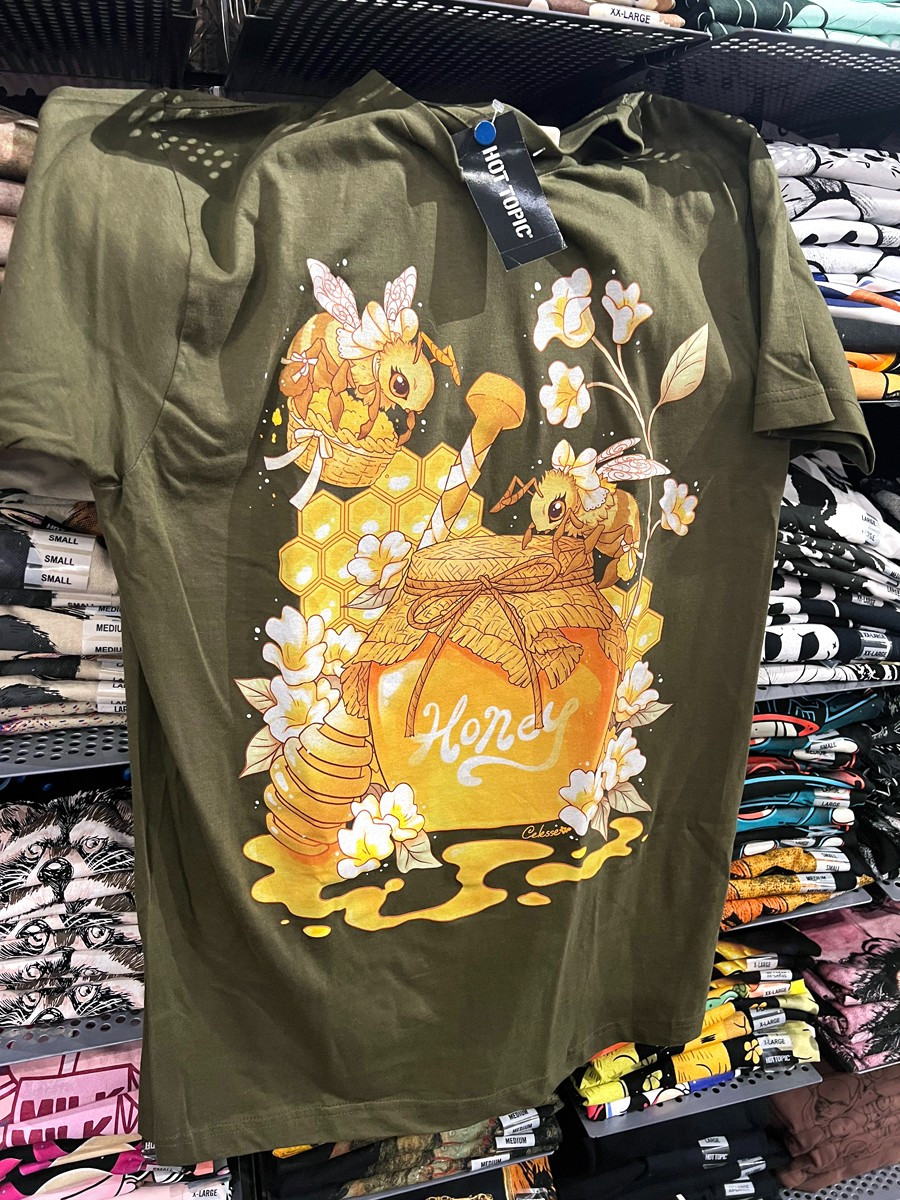 A photo of an olive green t-shirt hung on a wall of shelves of stacked shirts, the print featuring a jar of honey with pretty bees wearing white bows flying around it