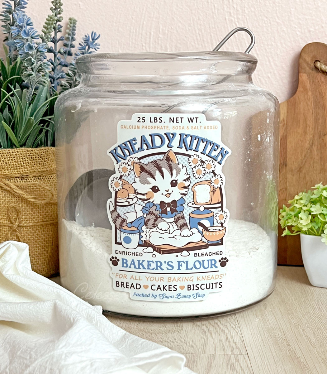 A photo of a large glass flour container on a counter with plants, a cutting board and a tea towel surrounding it. The container has a label sticker on it featuring a faux logo for Kneady Kitten Baker's Flour with a kitten in a frilly apron kneading dough on a cutting board