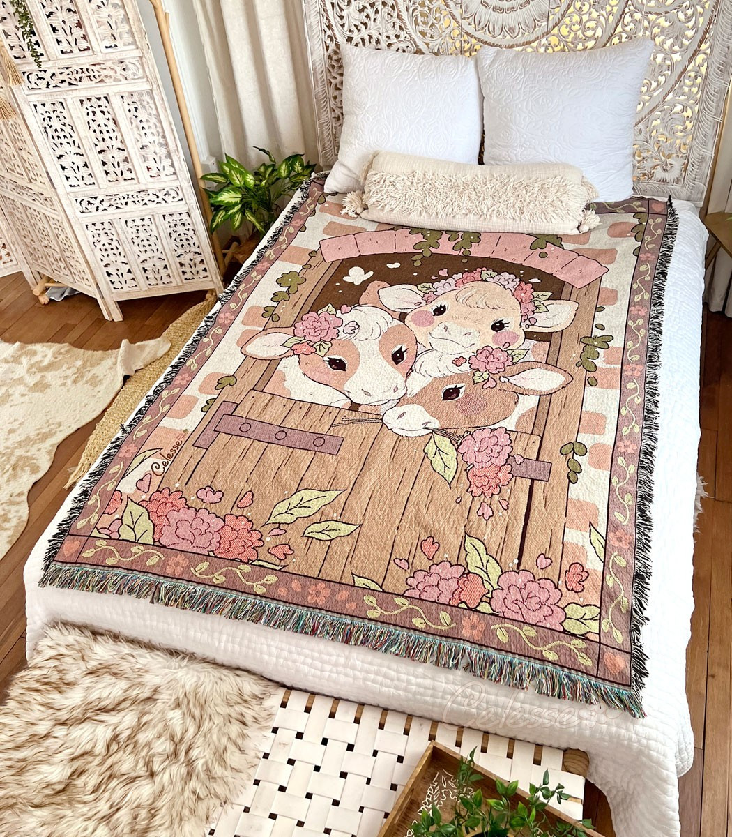 A photo of a woven blanket featuring an image of 3 country cows looking through a barn door set in a brick wall. The blanket is laid out on a bed in a soft white bedroom with dark wood flooring.