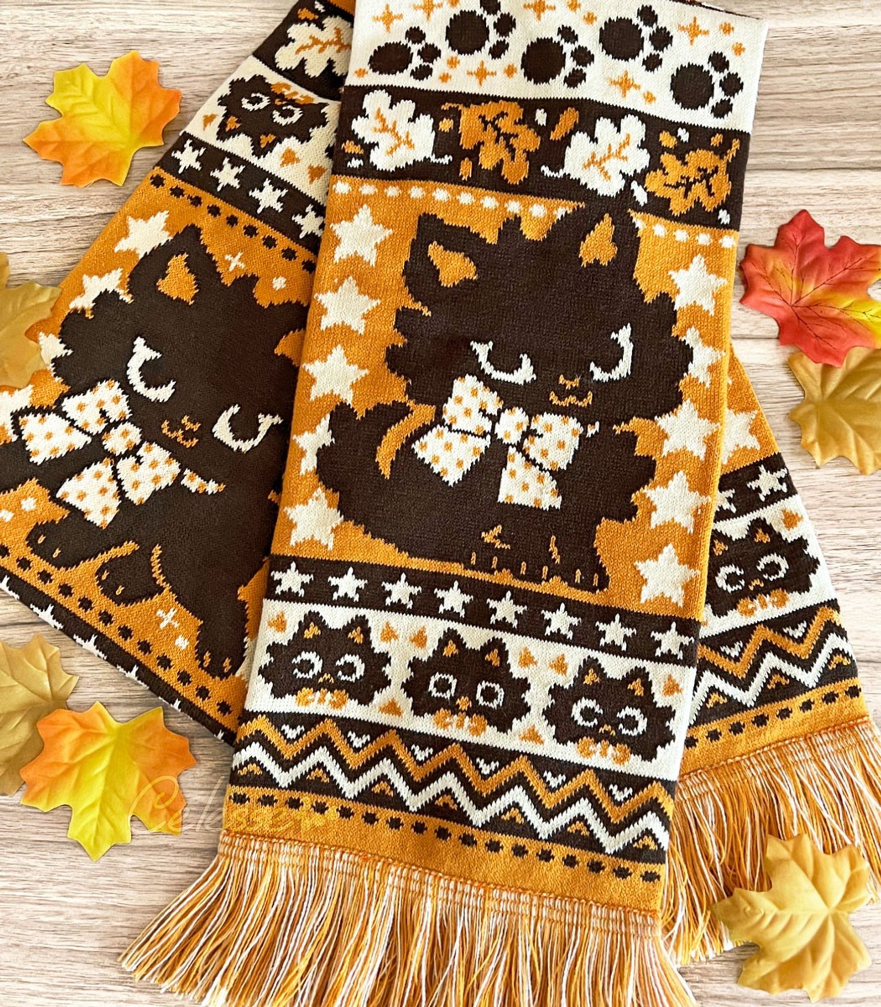 A close-up photo of a Halloween-themed knit scarf made of orange, dark brown and pale yellow yarn. A black kitten with a yellow polka dot bow poses amidst rows of stars, dots, leaves and other spooky motifs knit into the design.