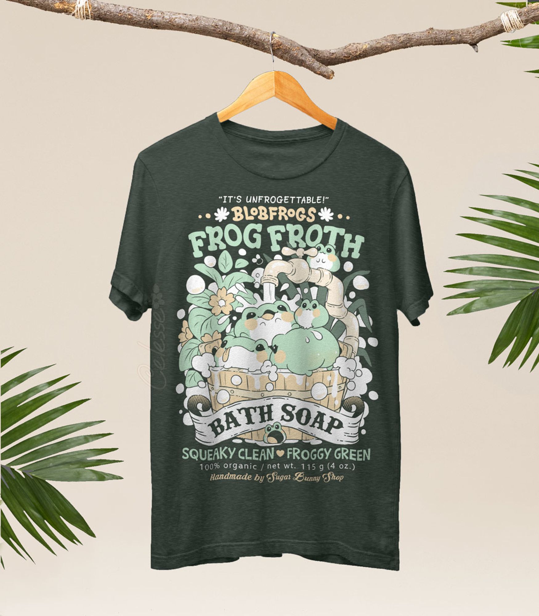 A photo of a heather forest green shirt on a wood hanger, hung from a branch with palm fronds on either side, the printed design features a vintage-style logo for Blobfrogs Frog Froth Bath Soap with a wood tub of fat squishy frogs taking a bubble bath