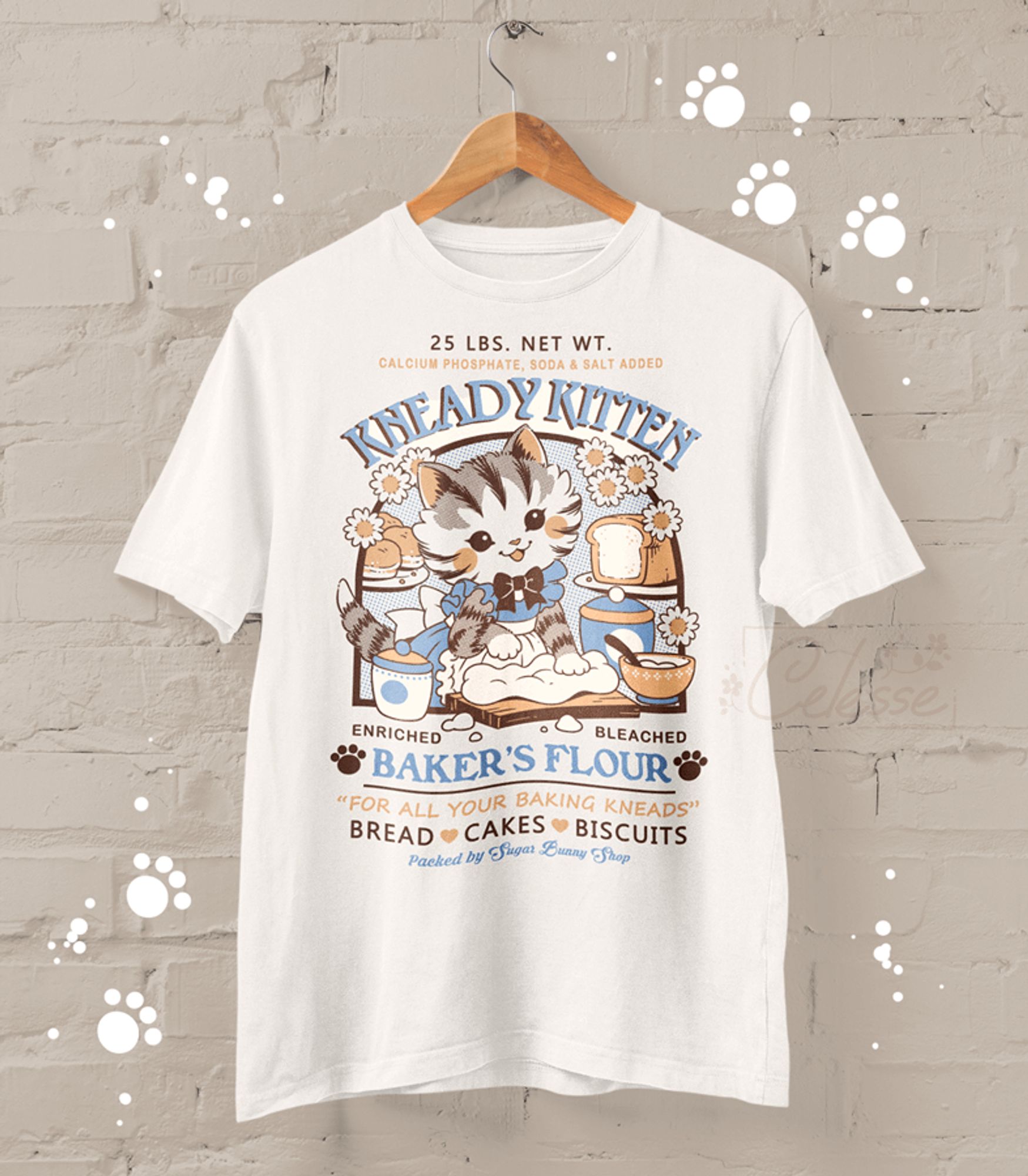 A photo of a shirt featuring an old-timey graphic design for Kneady Kitten Baker's Flour. The logo features a kitten in an apron kneading dough with bread, bowls and flowers around it. The text reads "For all your baking kneads - BREAD - CAKES - BISCUITS - Packed by Sugar Bunny Shop"