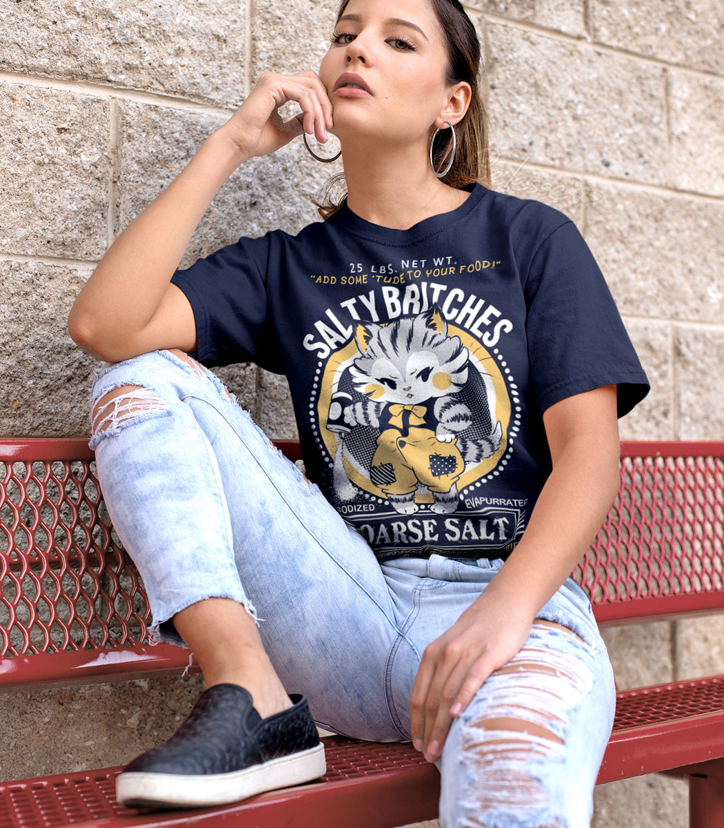 A young woman sitting on a bench with her foot  up on the seat and elbow rested on it, wearing a navy blue shirt with a faux logo for Salty Britches Coarse Salt. The logo features a grumpy kitten dumping a container of salt in a petulant manner