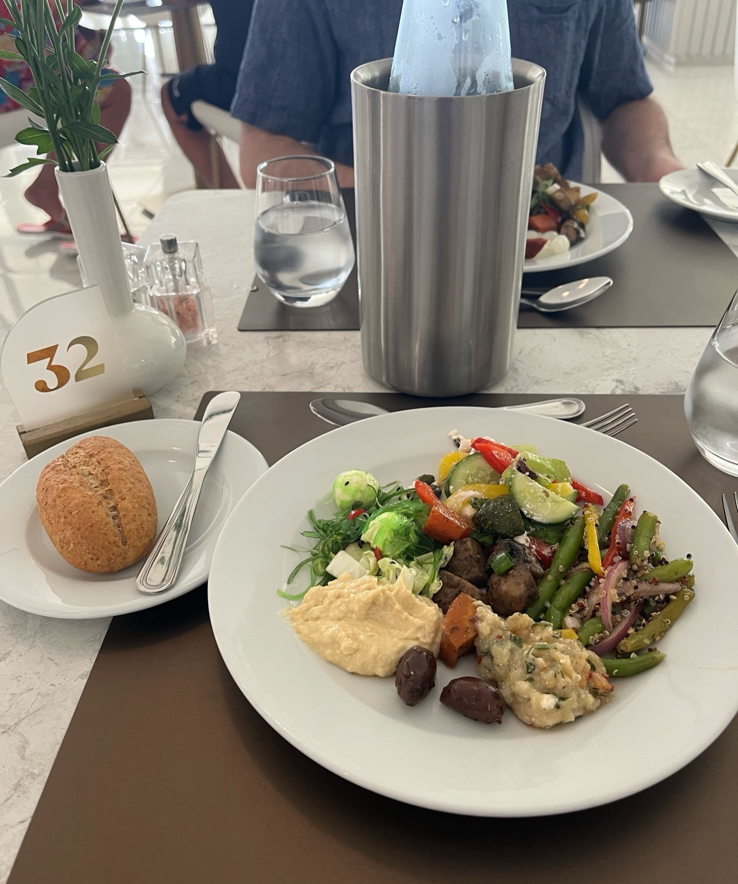 Een gedekte tafel met een bord vol verschillende kleurrijke salades. Ernaast een bordje met een broodje. En staat een karaf met een fles water en glazen water.