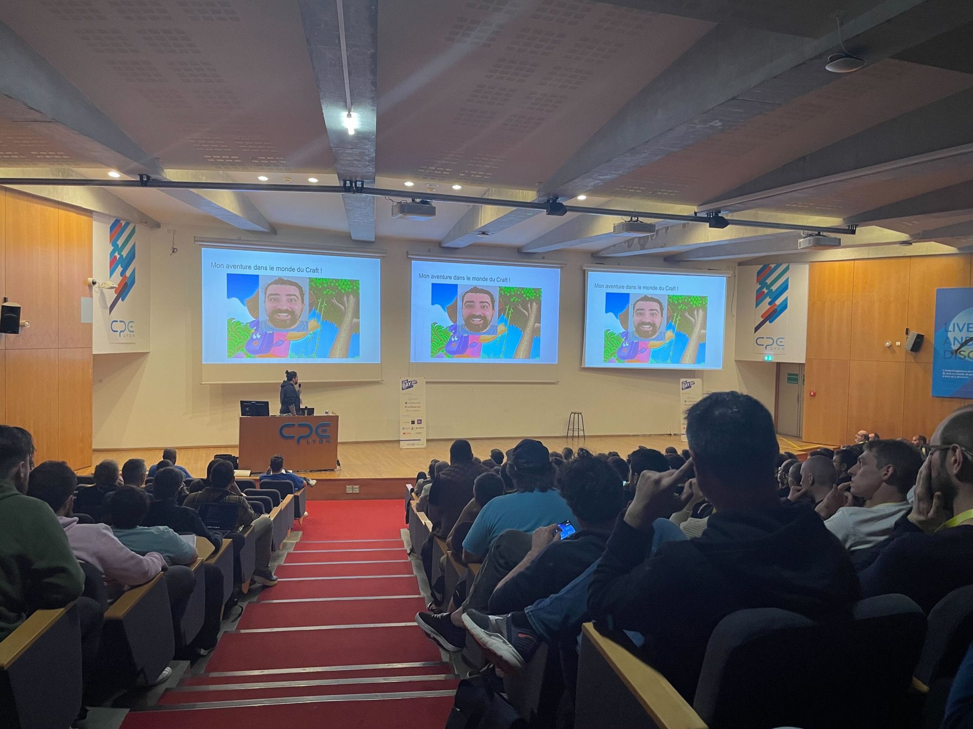 Public assis dans l'amphi de CPE à Lyon. Les slides du speaker sont sur 3 écrans géants, le speaker est sur scène. Photo par Nathan Boiron.