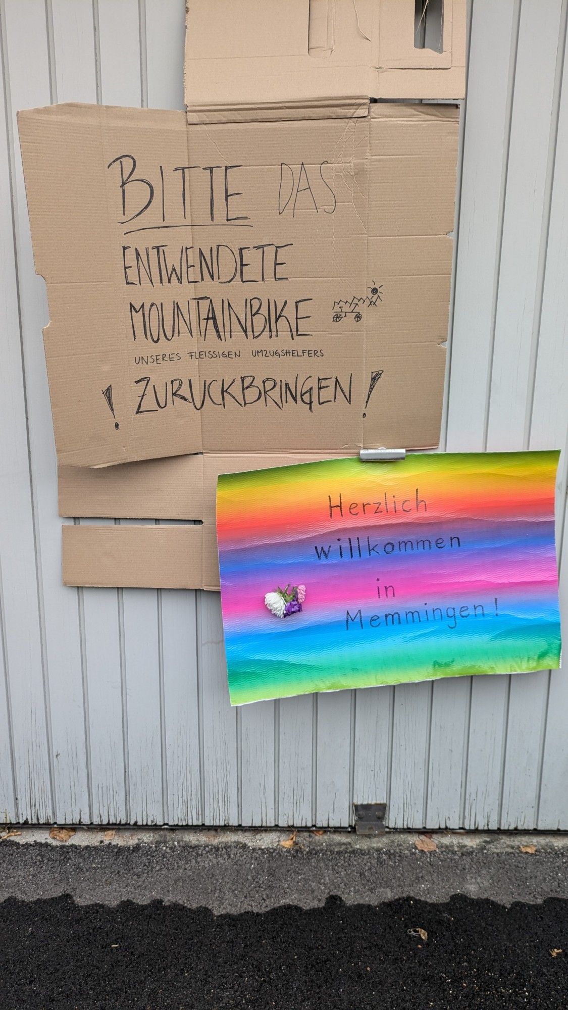 An einer Garage hängt ein, auf Karton geschriebener Aufruf: "BITTE das entwendete Mountainbike unseres fleißigen Umzughelfers !ZURÜCKBRINGEN!"

Darunter auf einem regenbogenfarbenen Karton: "Herzlich willkommen in Memmingen!"