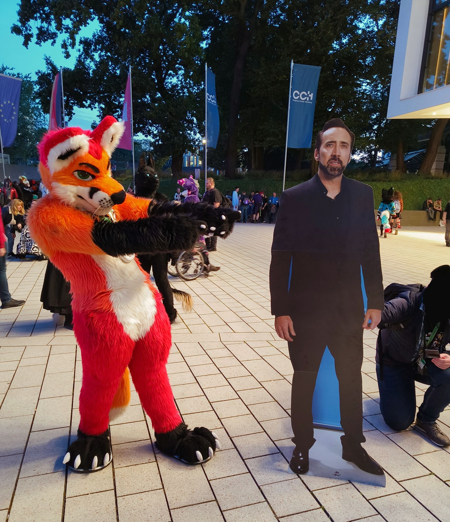 Fursuit photo of Loimu The Fox pointing at a life-size cardboard cutout of Nick Cage. Eurofurence con crowd fill the background scenery.