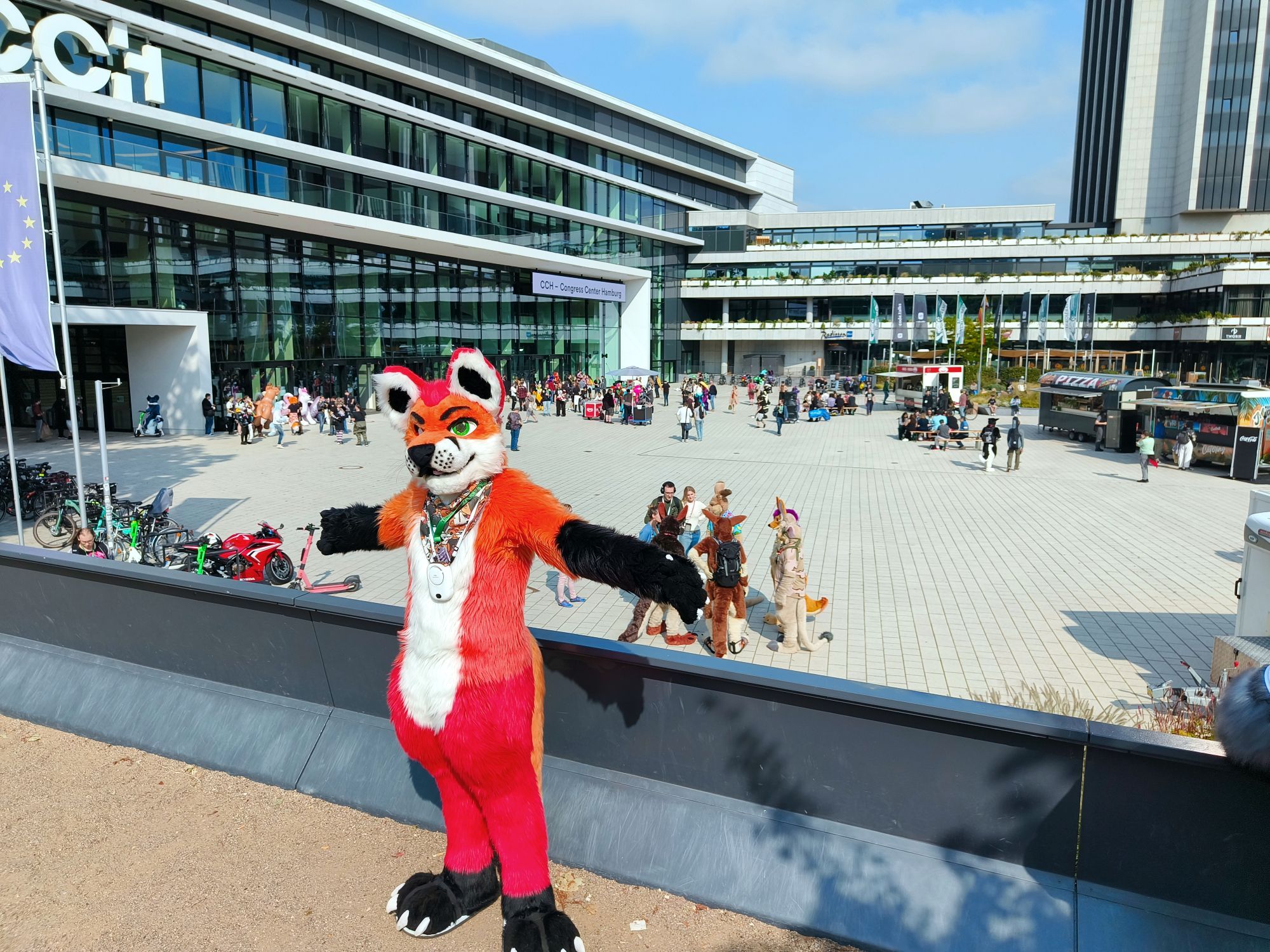 Fursuit photo of Loimu The Fox posing on a balcony above the front plaza of the Hamburg Conference Center where Eurofurence is being held. Lots of convention goers are spread out around the plaza behind.