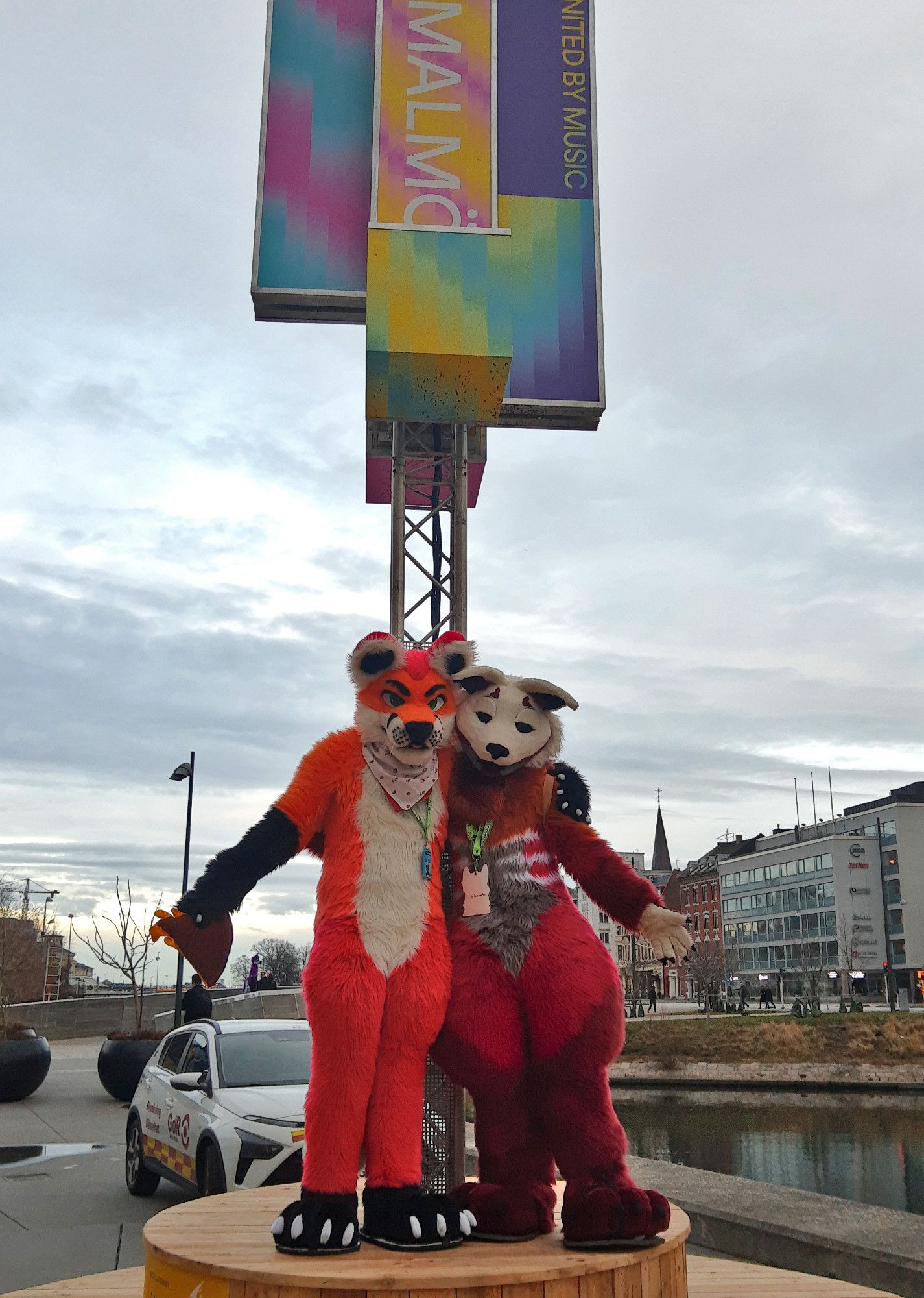 Fursuit photo of Loimu The Fox and who I presume to be a skulldog (but whose name I unfortunately forgot) standing on a pedestal with a Malmö sign behind them. A cityscape can be seen on the background.