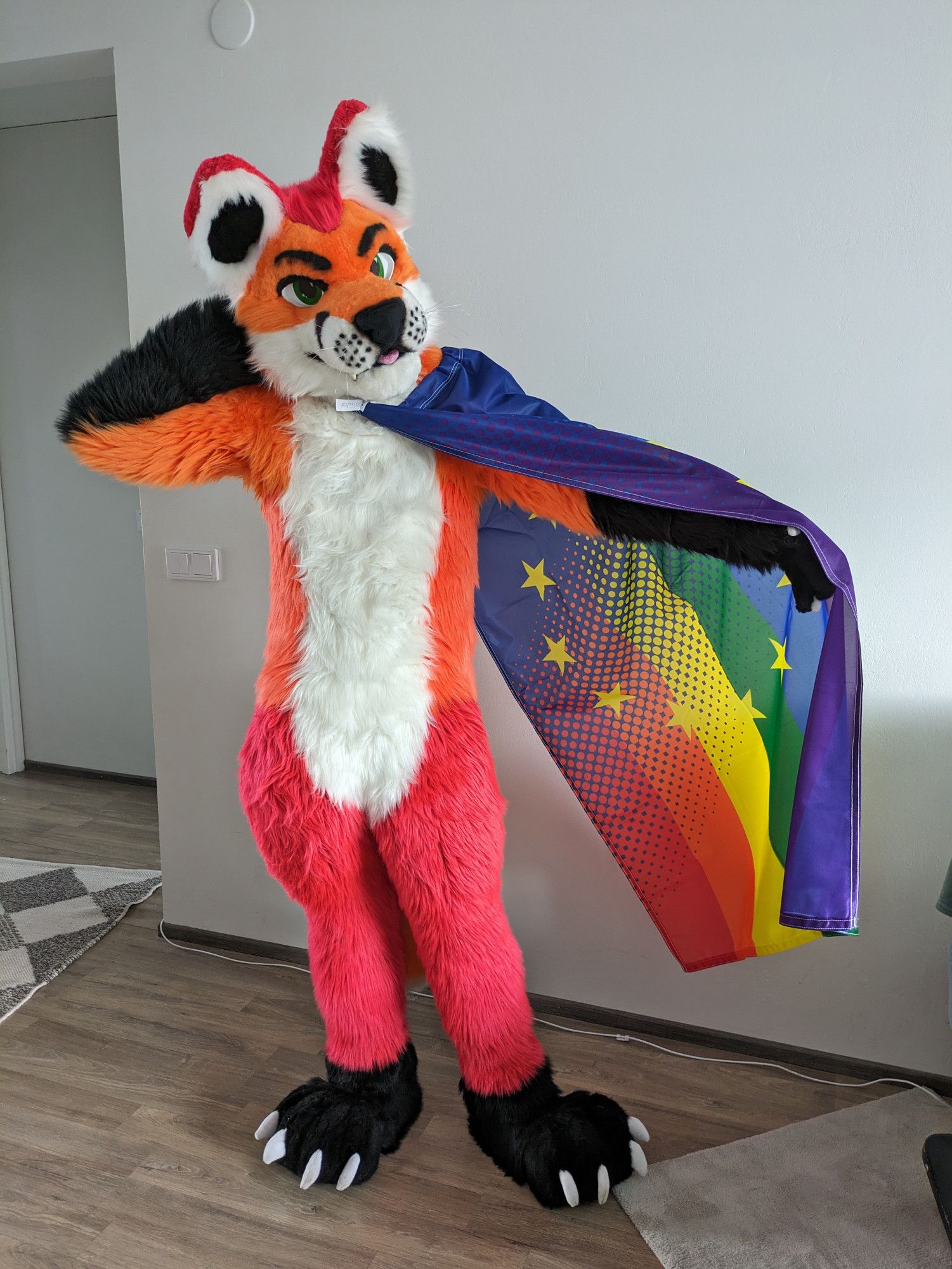 Fursuit photo of Loimu The Fox posing with a gradient rainbow-EU flag on his back.