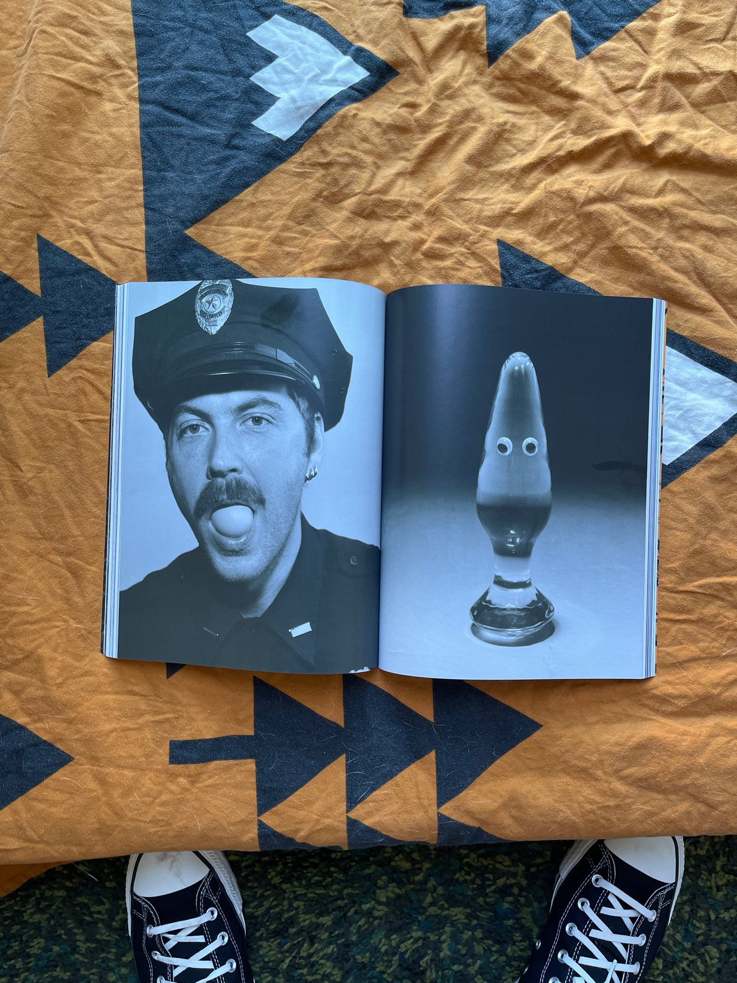 A magazine lying on an orange blanket opened to pages showing black and white photographs of a cop with an egg stuffed in his mouth and a butt plug with googly eyes.