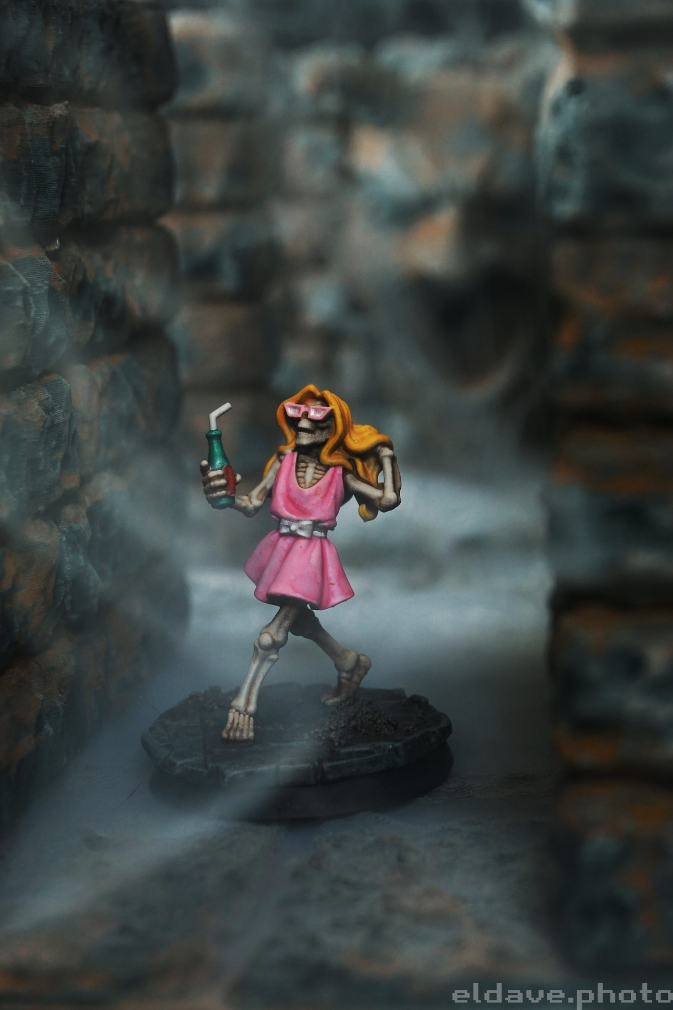 A skeleton walking out of a cold, gray stone dungeon wearing a pink sun dress and matching pink sunglasses. They are holding a green soda bottle with a straw in their right hand and running their left hand through a fake blonde wig. They are going to have an amazing vacation!