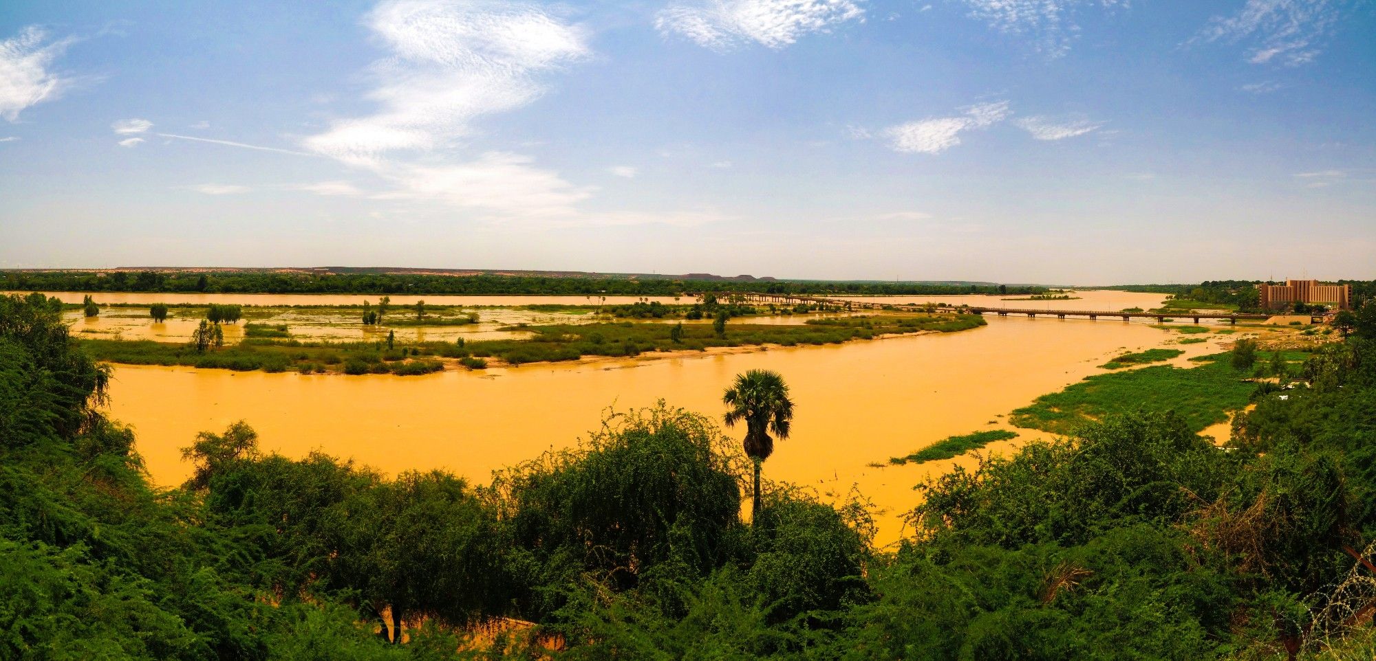 The Niger River flowing through a wide, flat floodplain, with muddy water flowing through many different side channels and a large bridge in the background