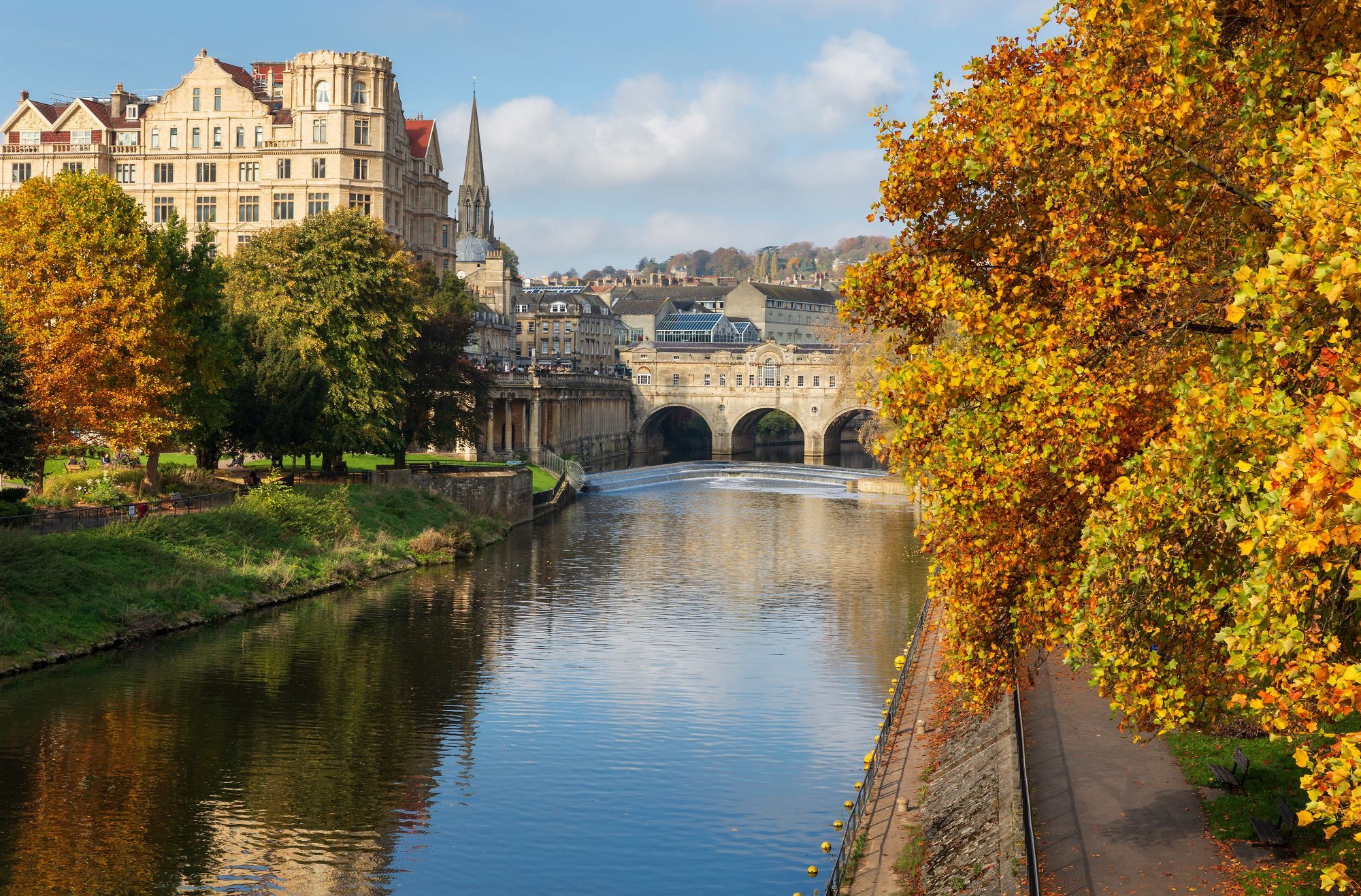 The large Avon river flowing between two well-developed banks, with yellow and green-leaved trees in the foreground and a large stone bridge crossing the river in the background, with a large building on the left bank and a city in the far background.