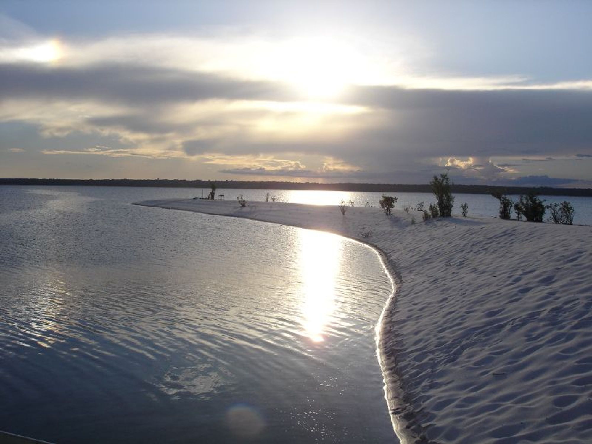 A river flowing along a white sandy bank that ends in the middle of the picture, showing where the North and South Para rivers converge with the sun falling behind clouds in the background