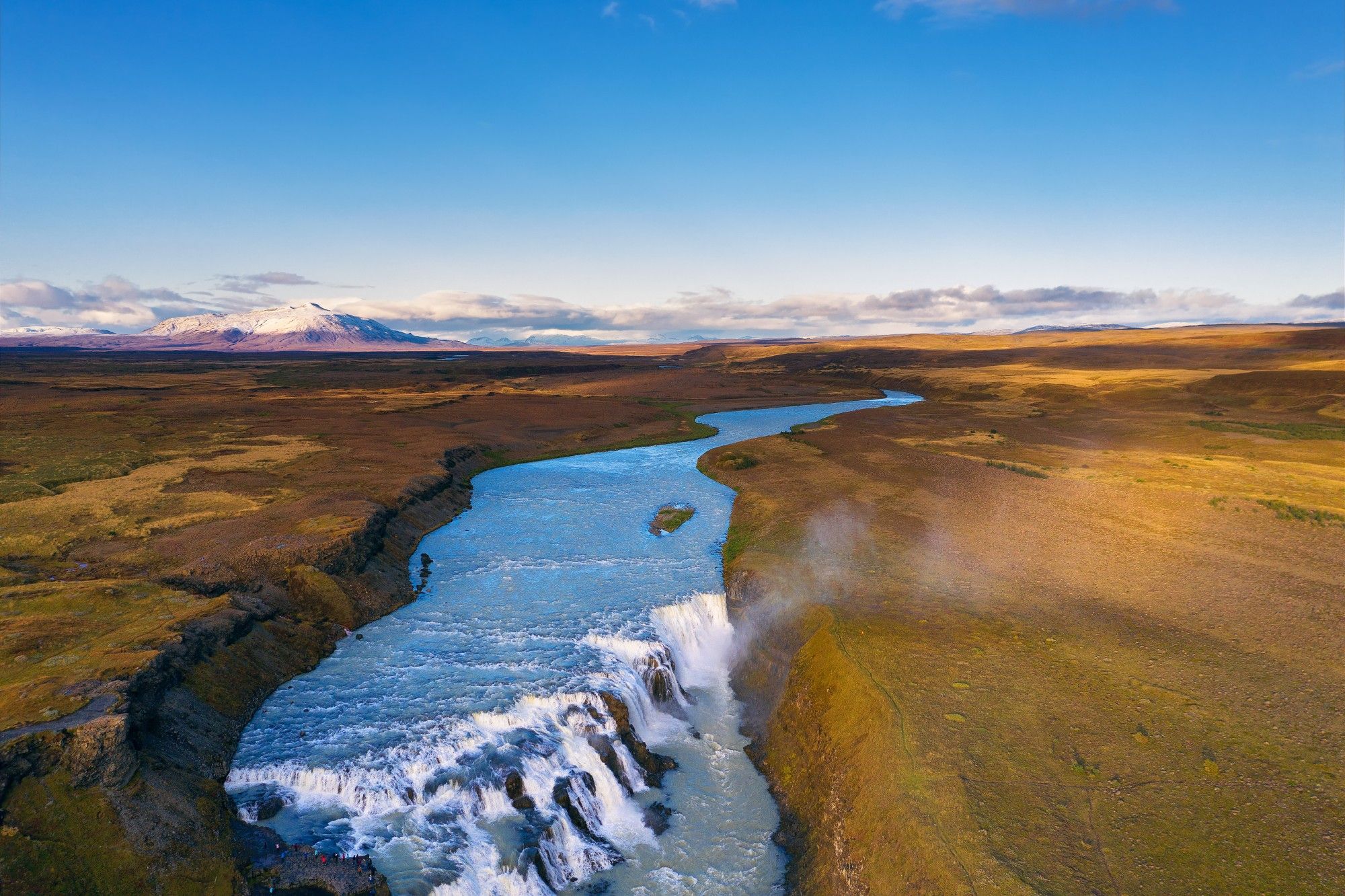 The Olfusa river as it flows through a wide plain before dropping through the famous Gullfoss waterfall, with bright shrubby vegetation on the riverbanks and a large mountain far in the background