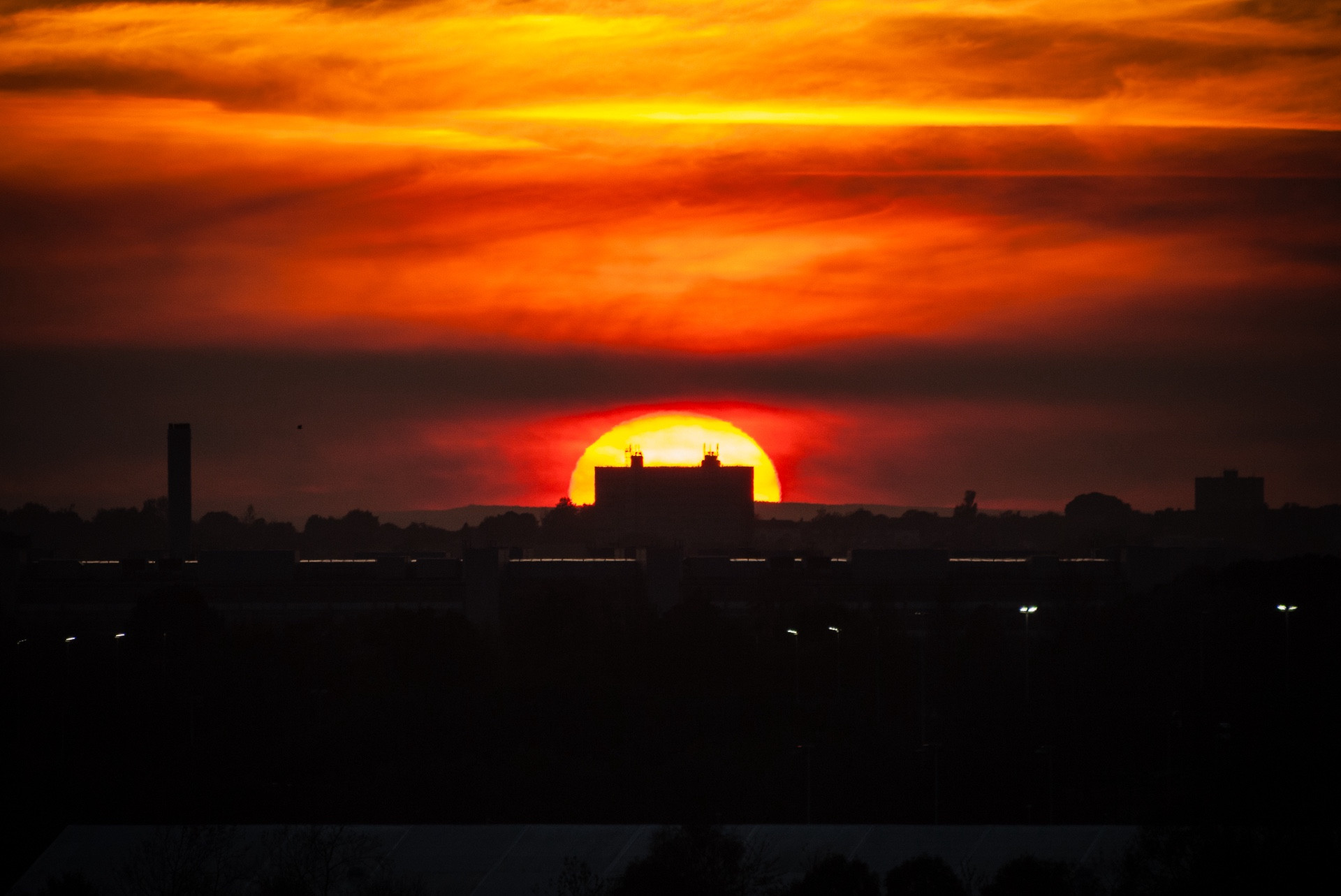 Colour photograph of the sun setting over Newcastle upon Tyne. The broiling clouds are rich red and orange. A distant building is perfectly and symmetrically silhouetted by the huge egg yolk setting sun.