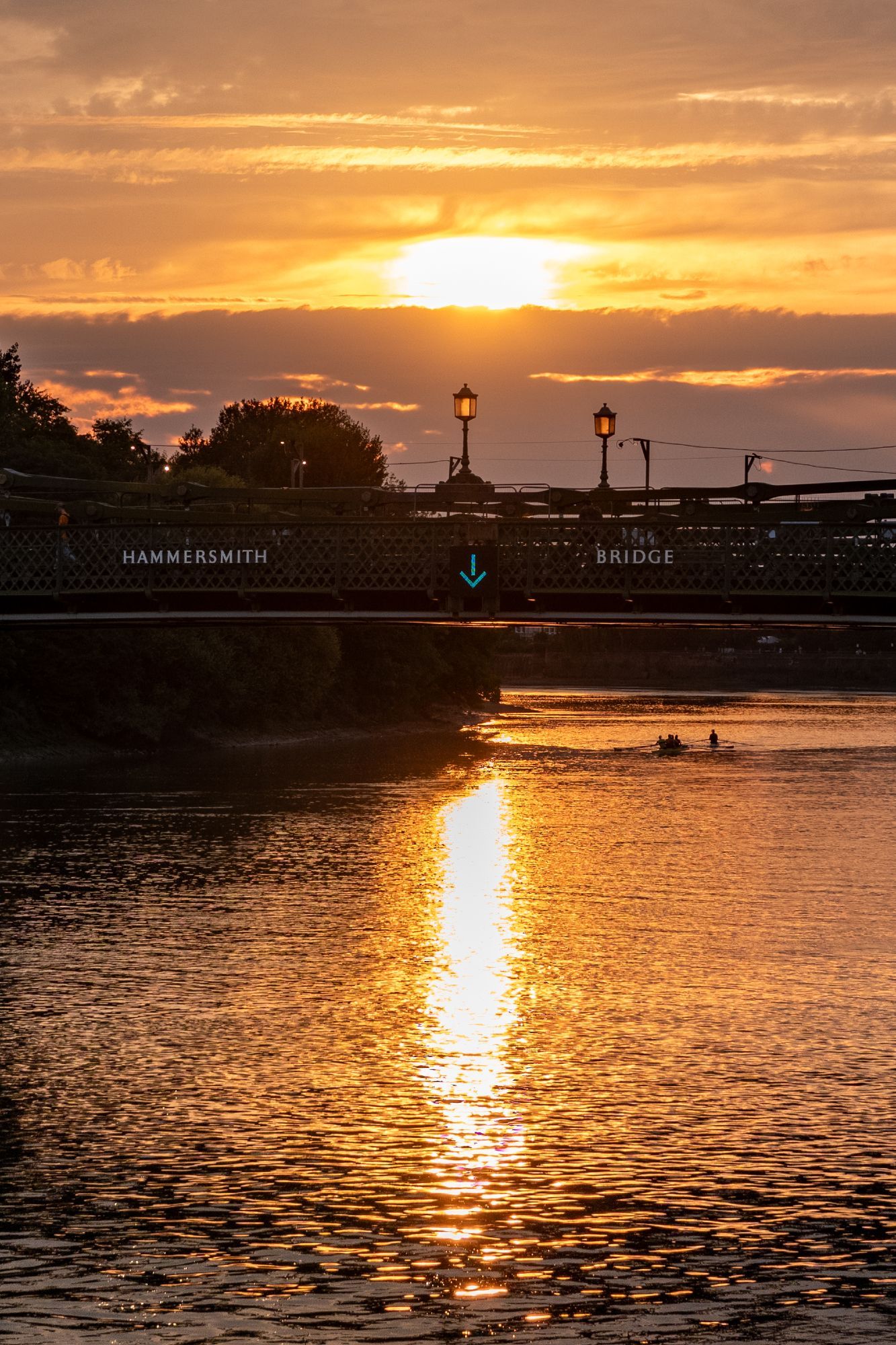 A sunset view of Hammersmith Bridge, the sun is disappearing into clouds, beneath that there's an arrow pointing down on the bridge, and directly beneath that is the sun reflected in a long line on the river. In the distance there's two rowboats