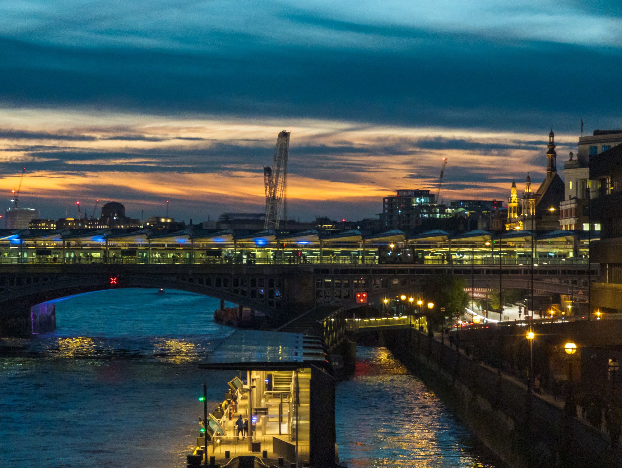 Sunset view of a railway bridge crossing a wide river, with a station on the bridge, amid a city.